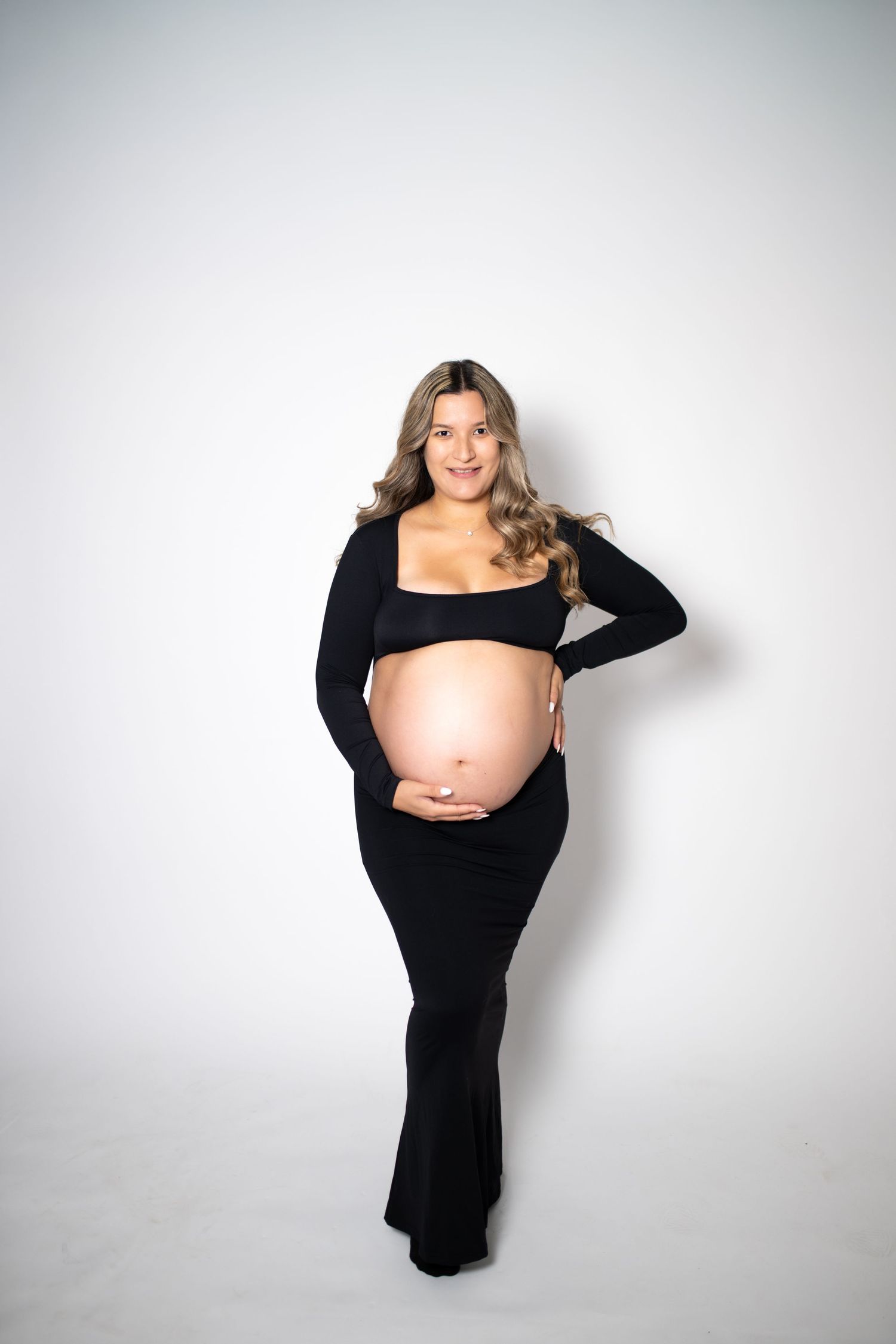 Pregnant woman in black crop top and skirt cradling baby bump, smiling at camera against white background.