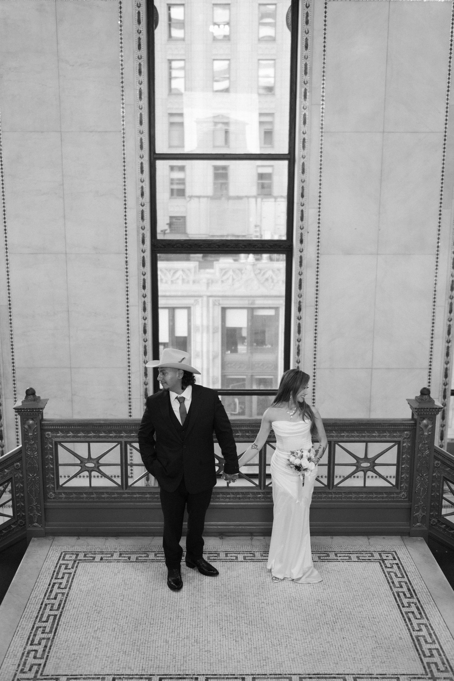 A couple in wedding attire stands in a grand room with tall windows and patterned floor.