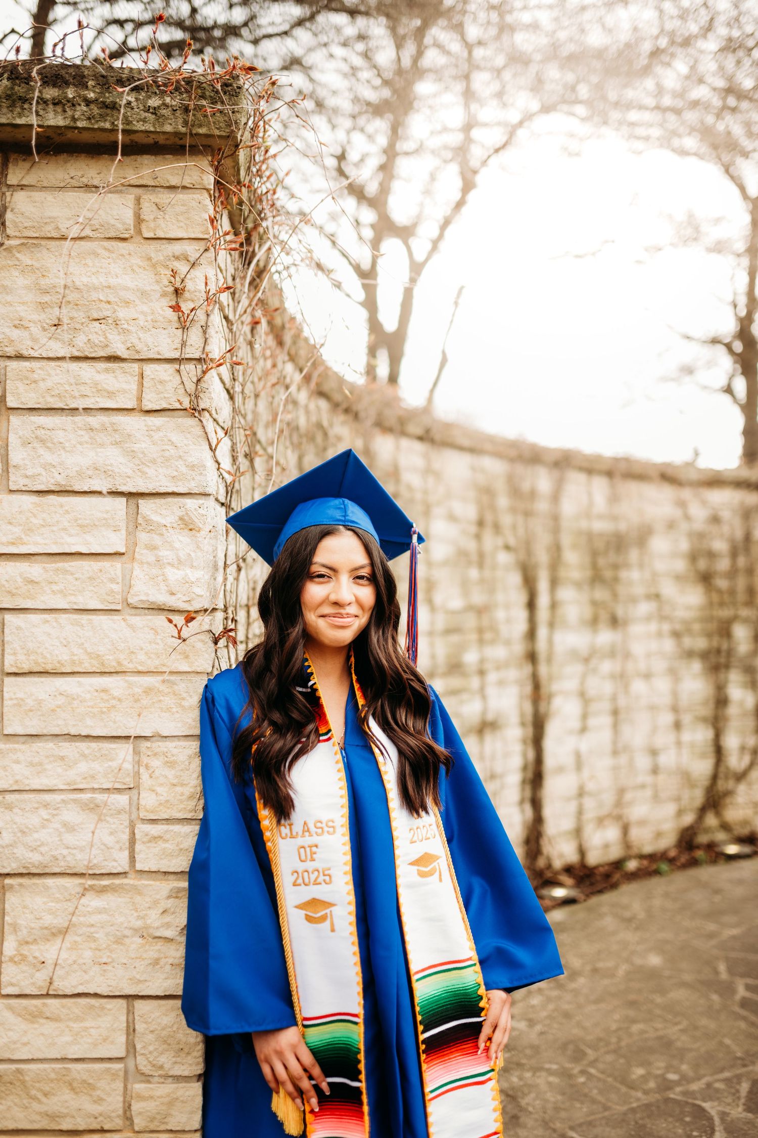 Graduate wearing blue cap and gown with colorful stole poses against stone wall.