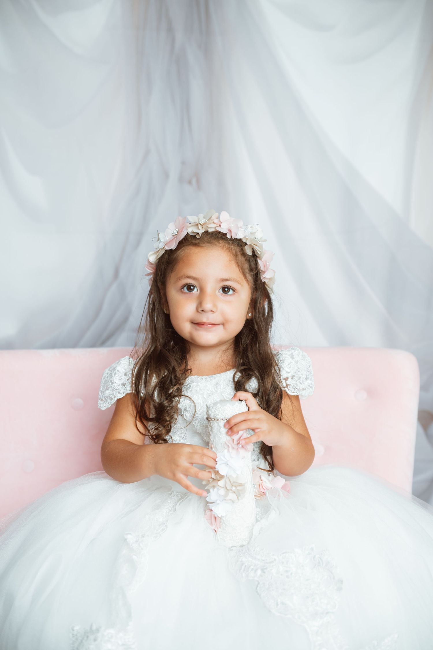 Child in ornate white dress and floral crown sitting against pink and white backdrop.