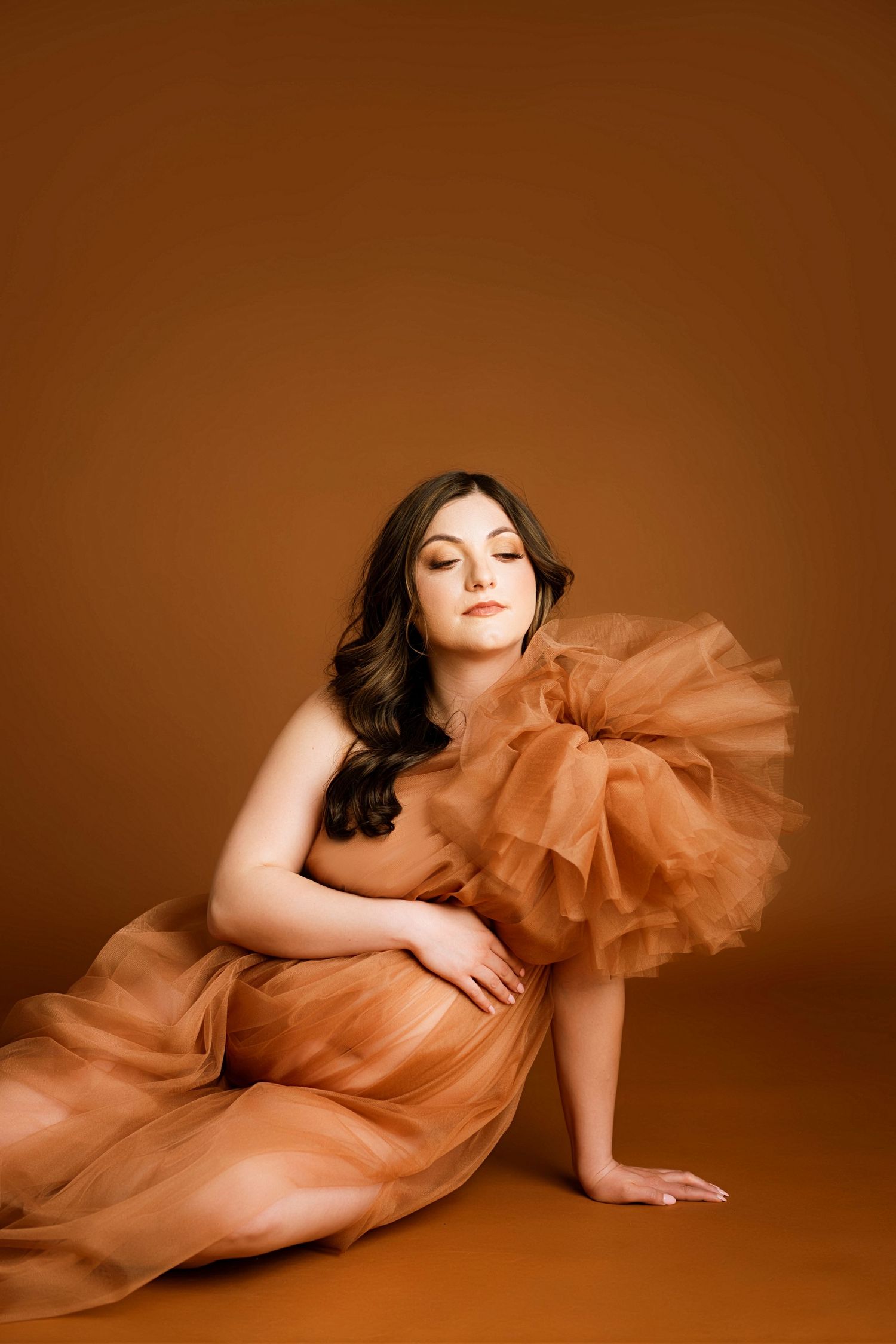 Woman in flowing rust-orange tulle gown sitting gracefully on studio floor against warm brown background.