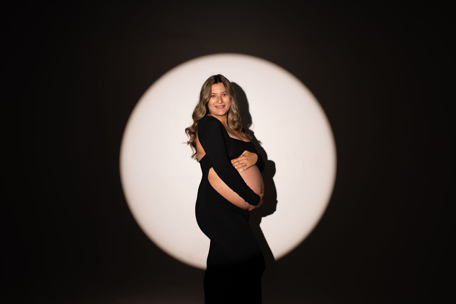 Pregnant woman in black dress poses elegantly against circular light backdrop in professional maternity portrait.