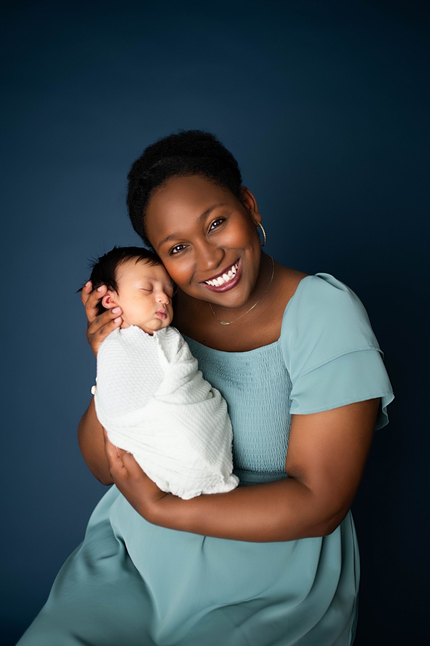 Smiling Black mother in teal dress holds sleeping swaddled newborn baby against dark blue background.