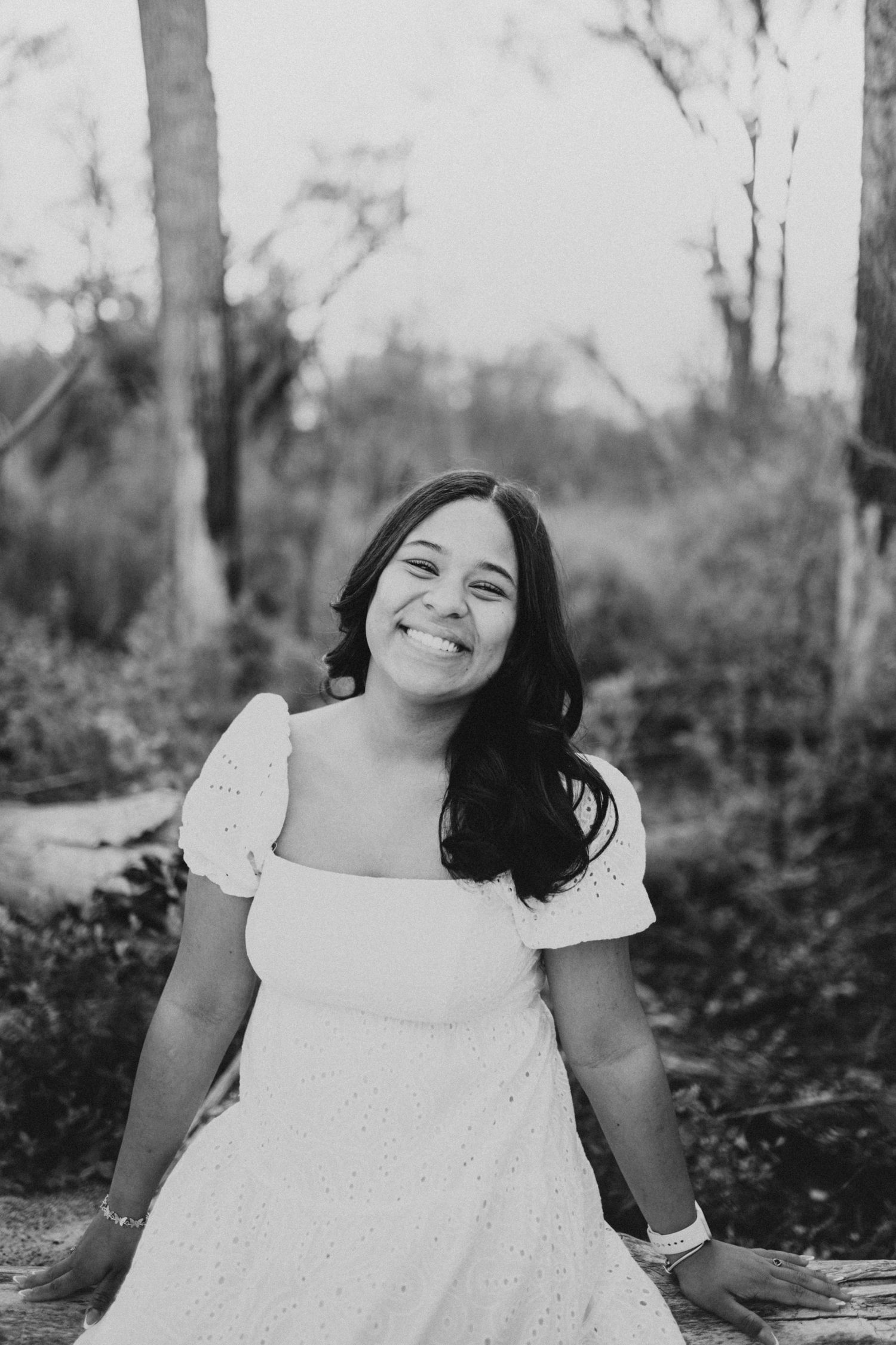 Graduate in white eyelet dress poses on log during outdoor photo session.