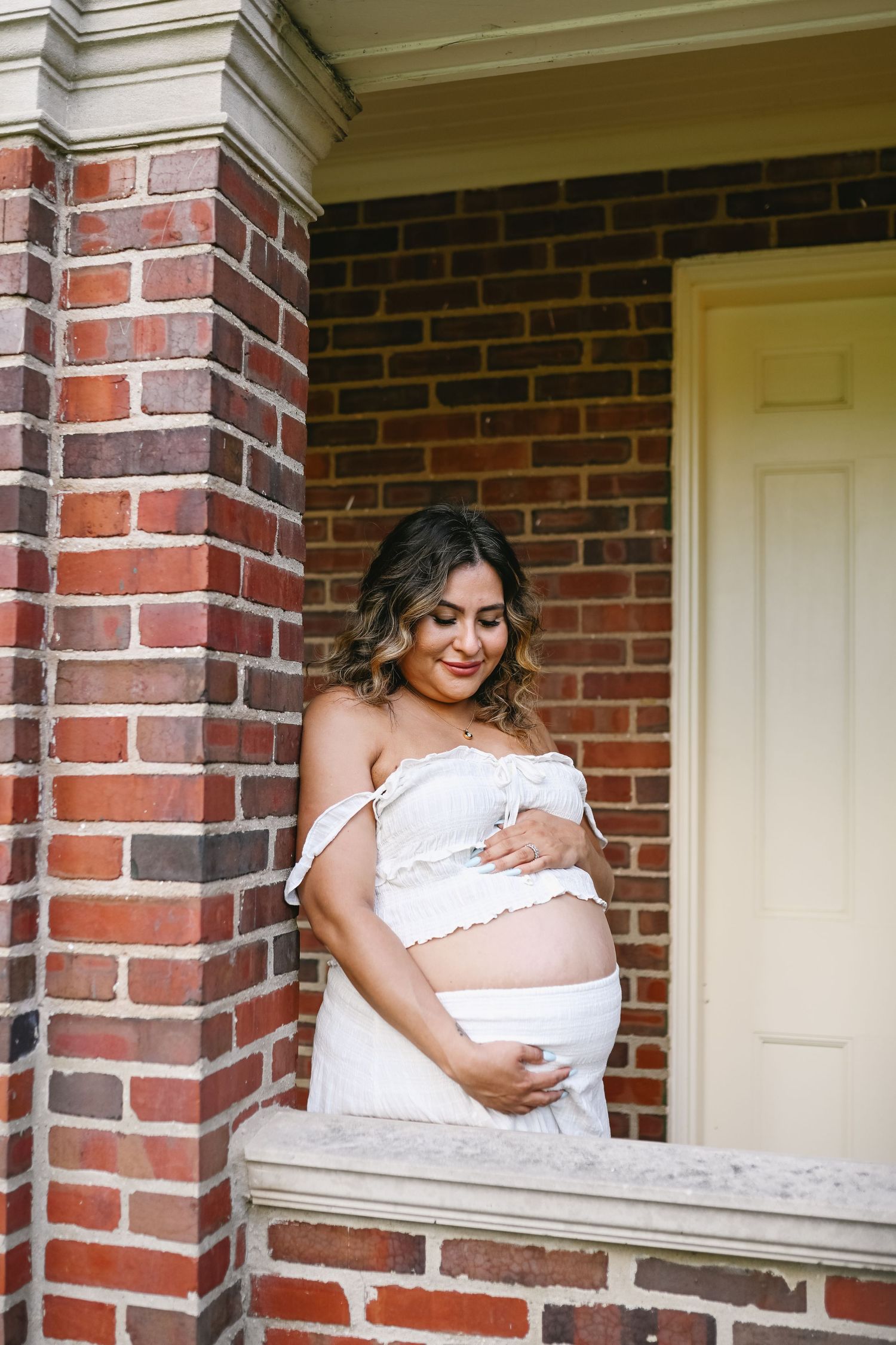 Pregnant woman in white off-shoulder maternity outfit cradling her baby bump, standing by brick wall.