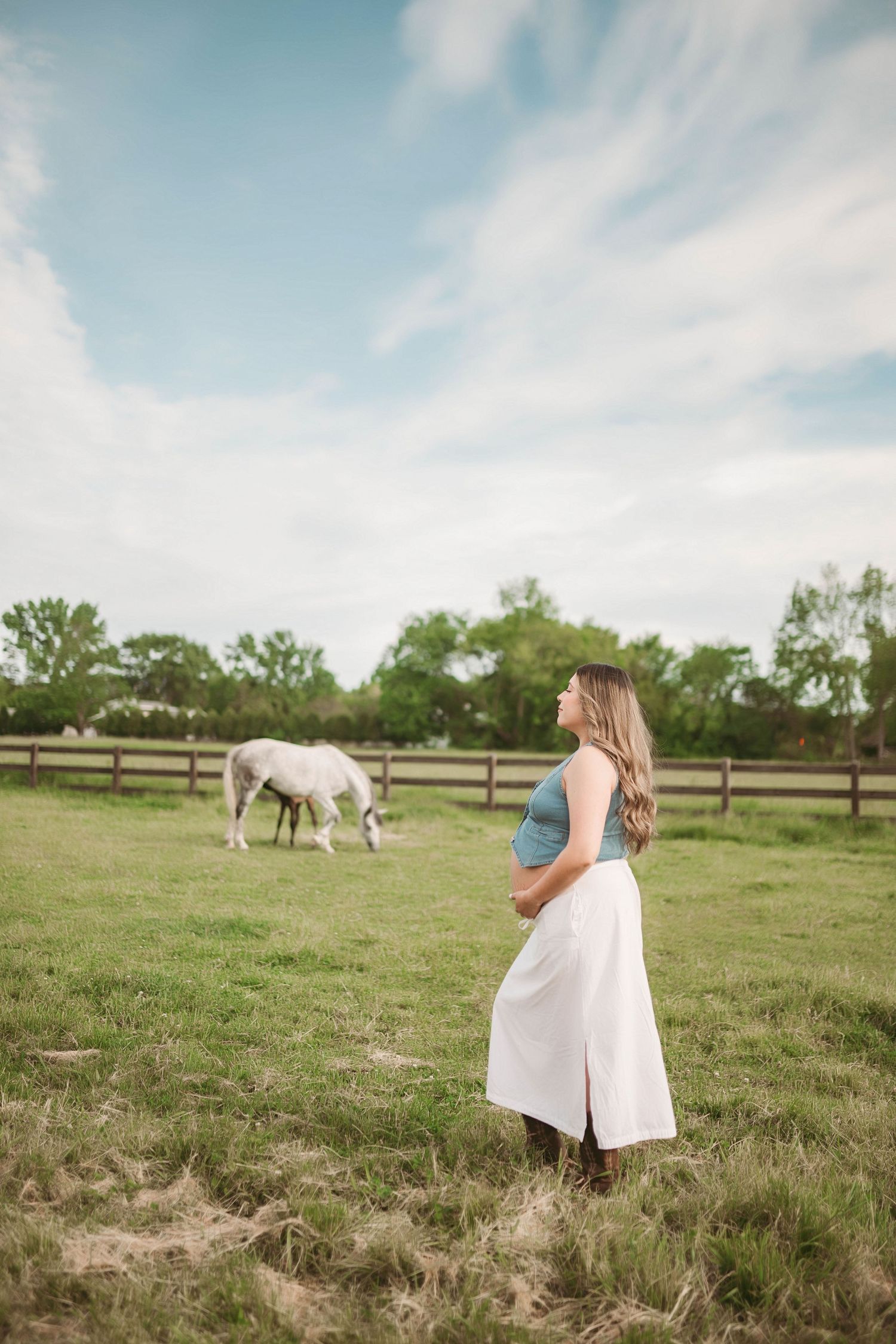 Pregnant woman in blue top and white skirt stands in green pasture with white horse grazing under cloudy sky.