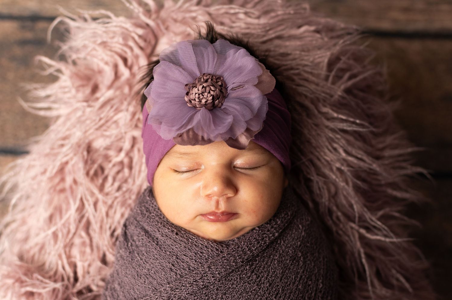 Sleeping newborn baby wrapped in purple knit swaddle with floral headband on fluffy pink fur backdrop.