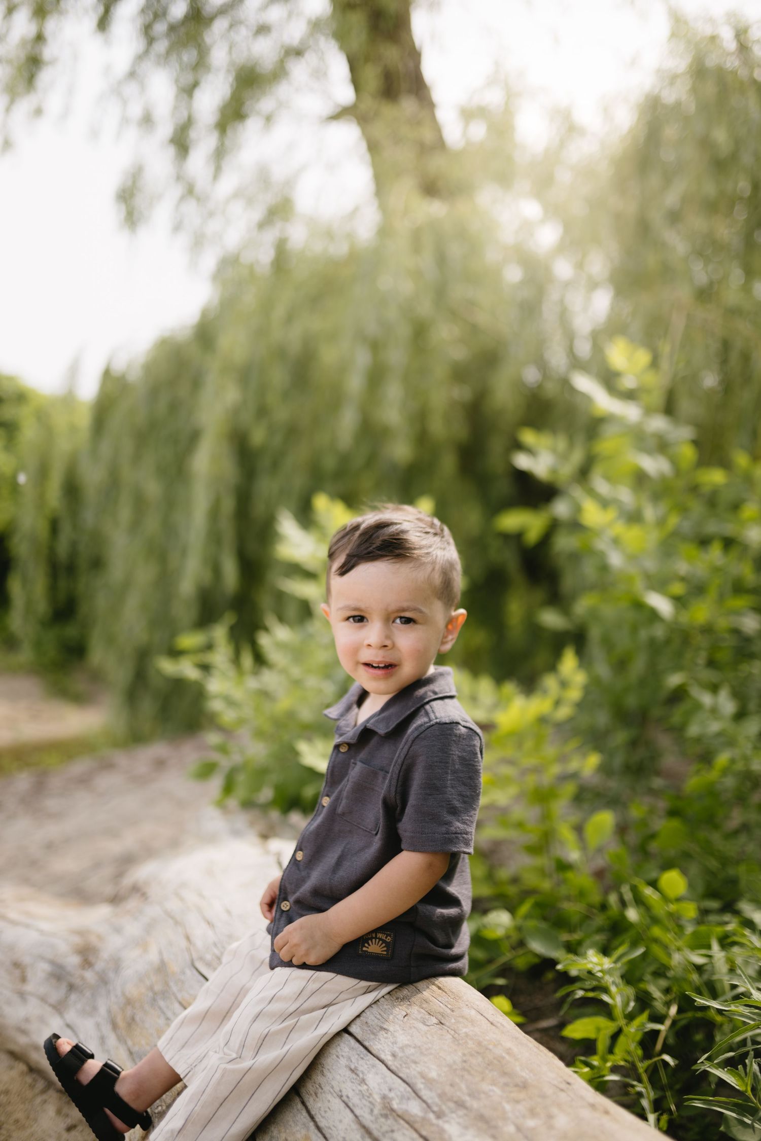 Young child sitting on fallen log surrounded by willow trees and greenery.