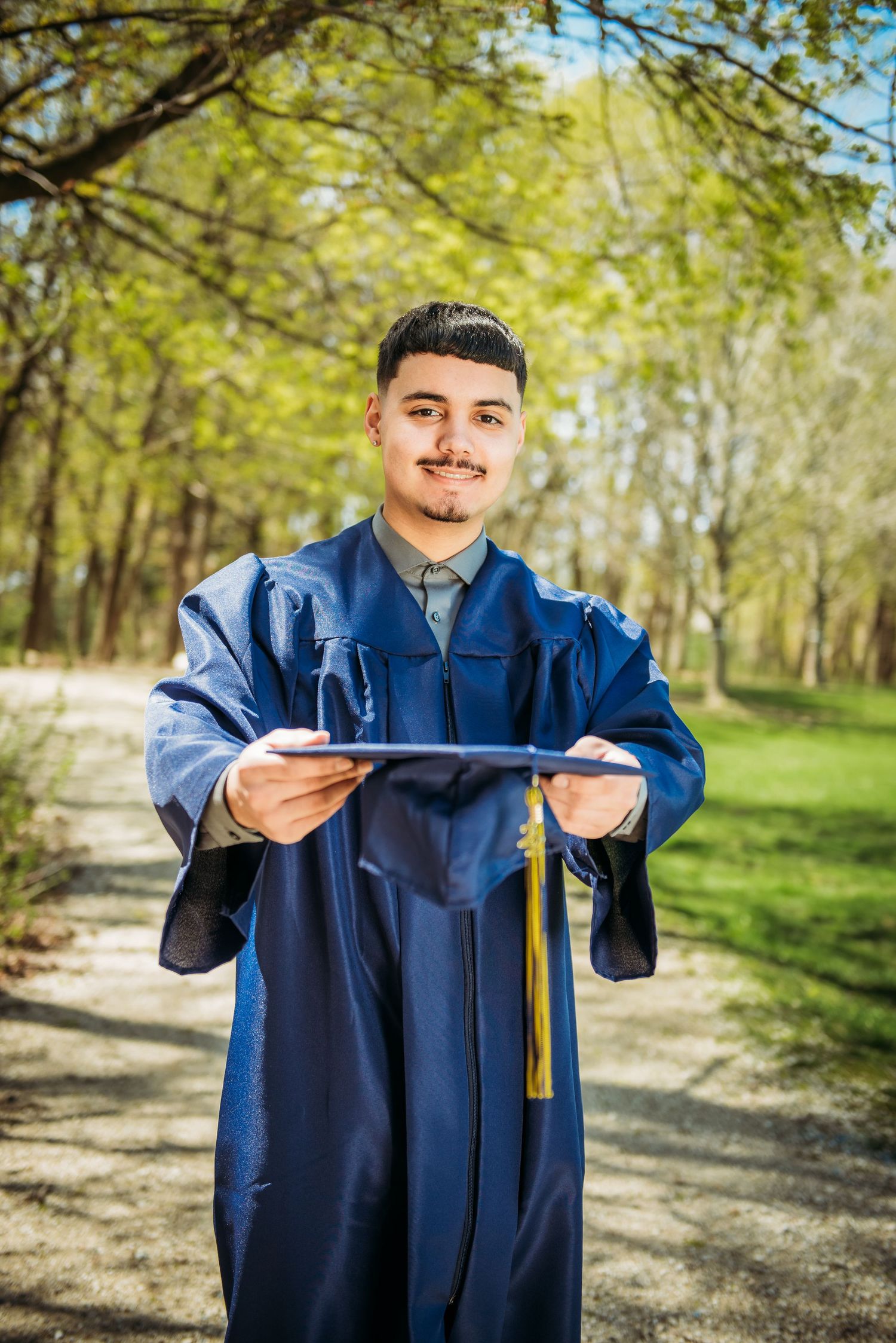 A graduate in blue cap and gown presents their diploma on a tree-lined path.