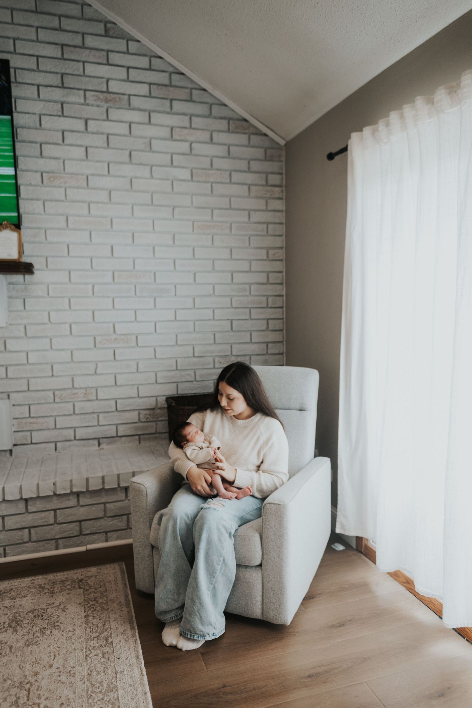 Young mother sitting in gray armchair holding newborn baby in cozy modern living room with brick wall.