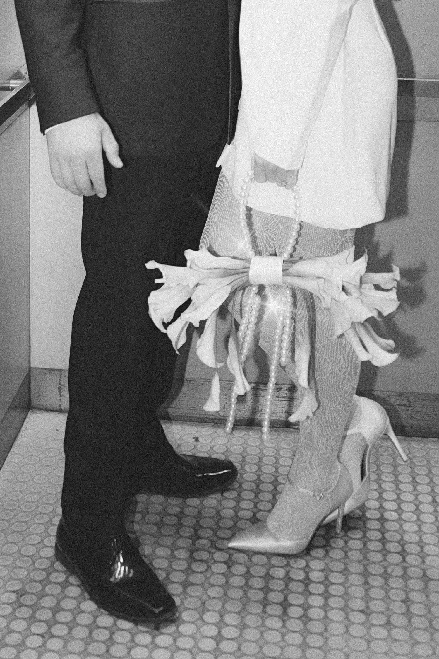 Black and white photo showing formal dress shoes and white heels on a tiled floor with a white gown and dark suit visible.