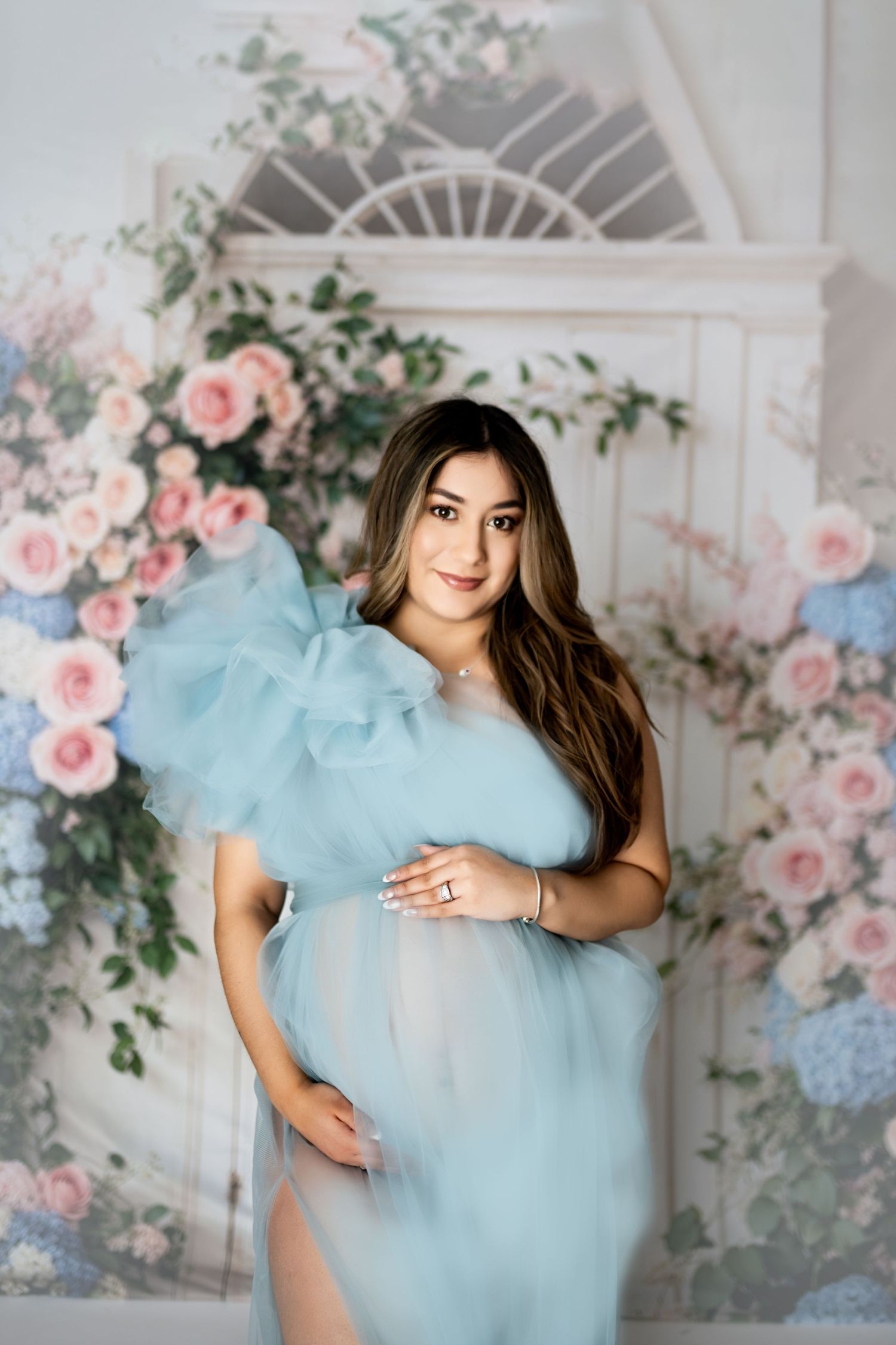 A woman in a light blue tulle dress poses against a floral backdrop in a studio setting.
