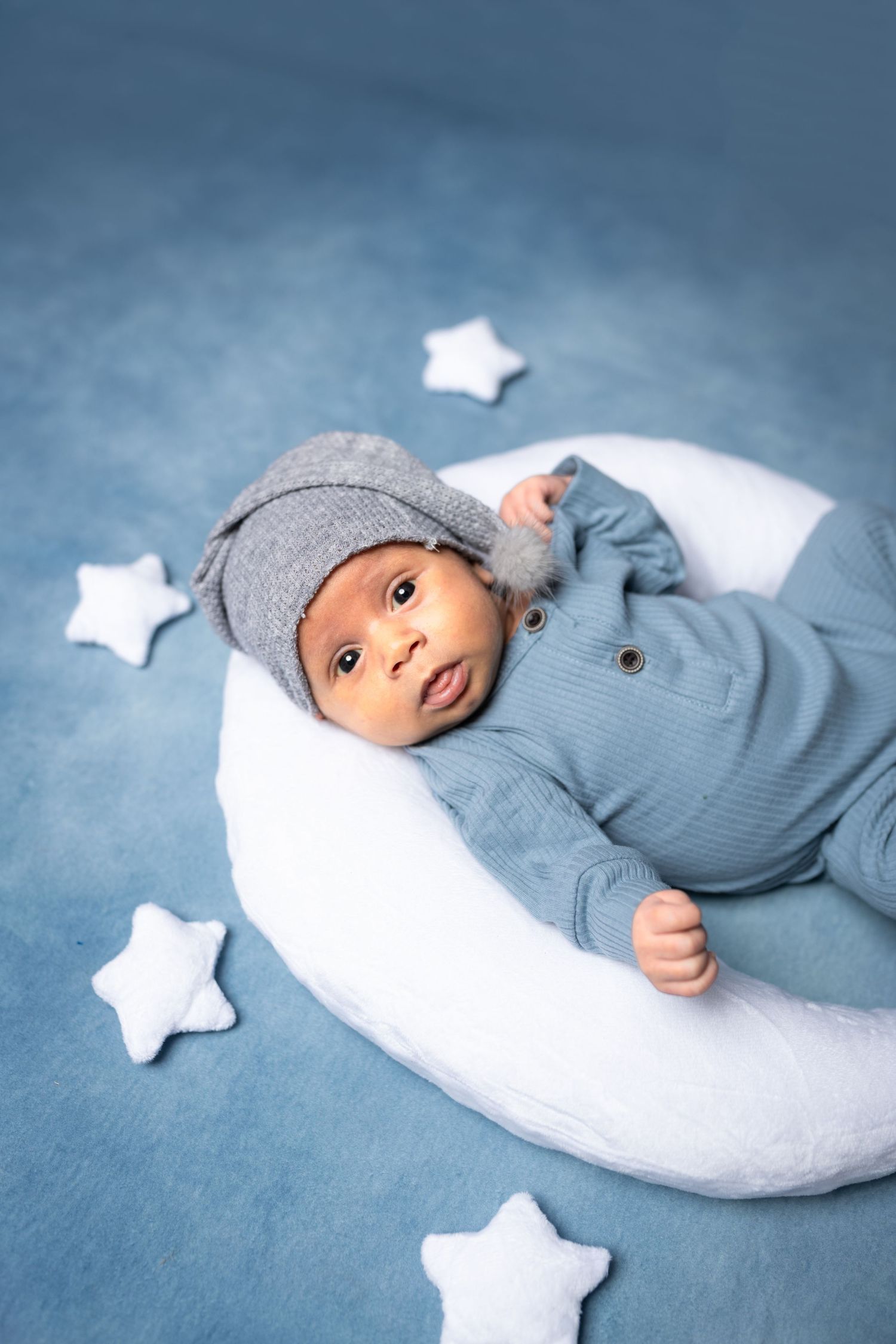 Newborn baby in blue outfit and gray knit hat resting on white moon pillow surrounded by star props.