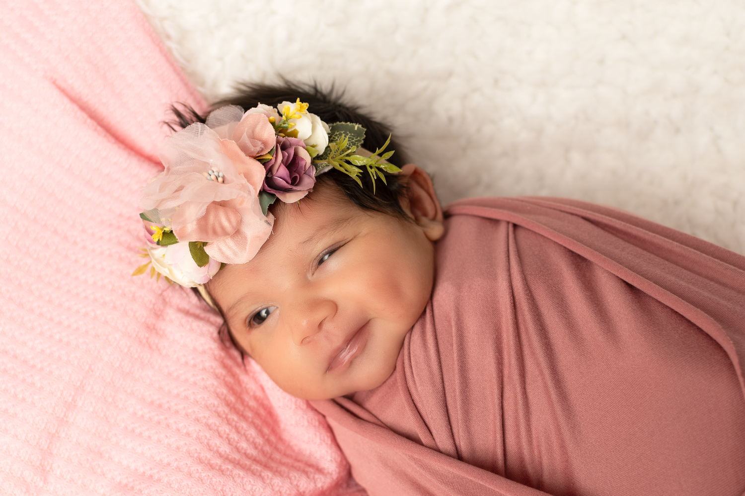 Newborn baby girl swaddled in pink wrap wearing a floral crown, smiling peacefully on a white fur blanket.