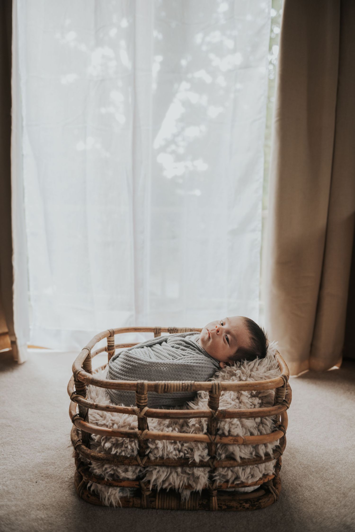 Newborn baby swaddled in gray knit blanket sleeping peacefully in a rustic wicker basket with fluffy fur lining.