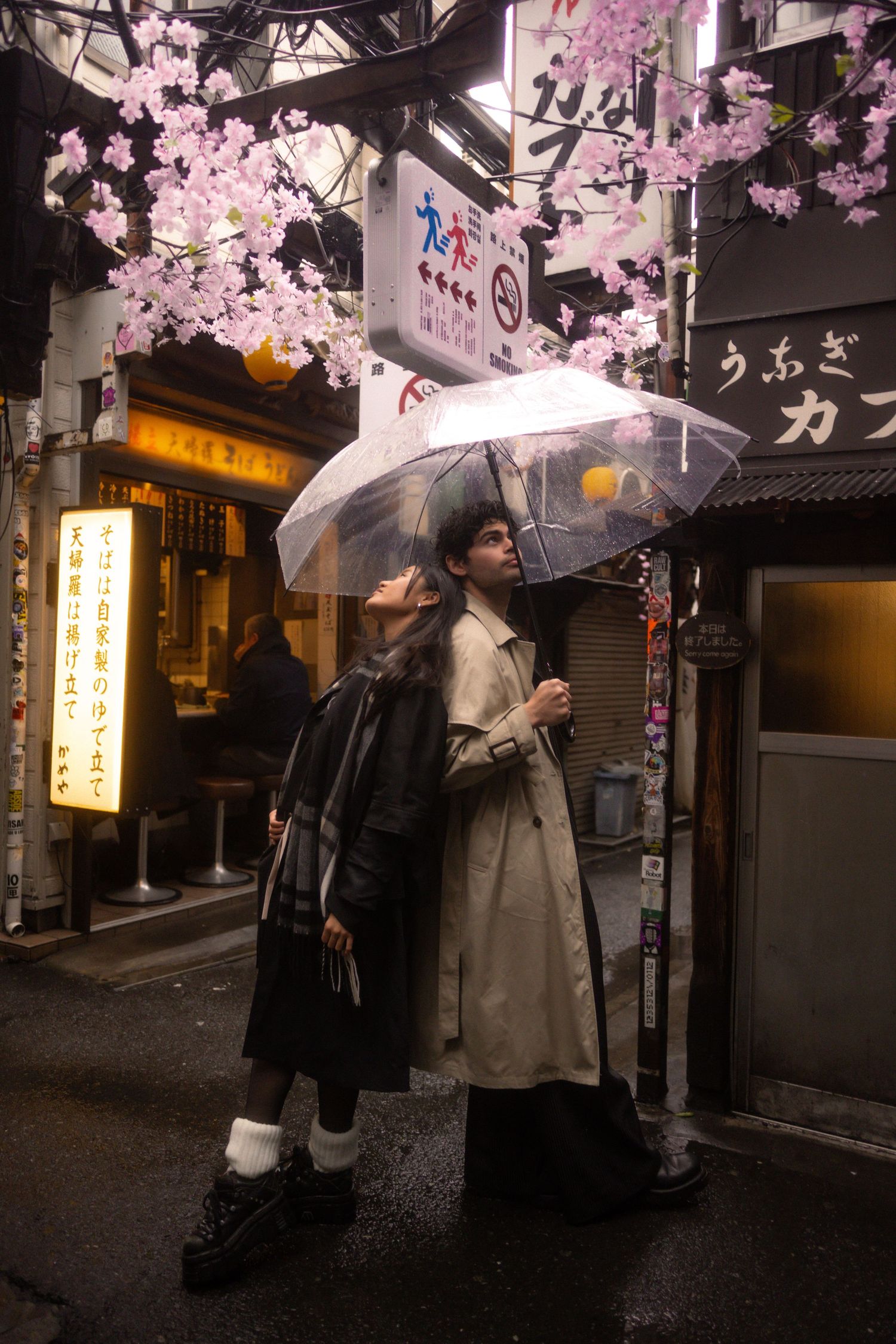 Two people with umbrellas stand beneath pink cherry blossoms on a rainy evening in a Japanese street with storefronts.