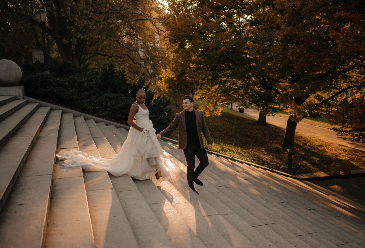 A wedding couple walks up concrete steps surrounded by autumn trees during golden hour sunset.