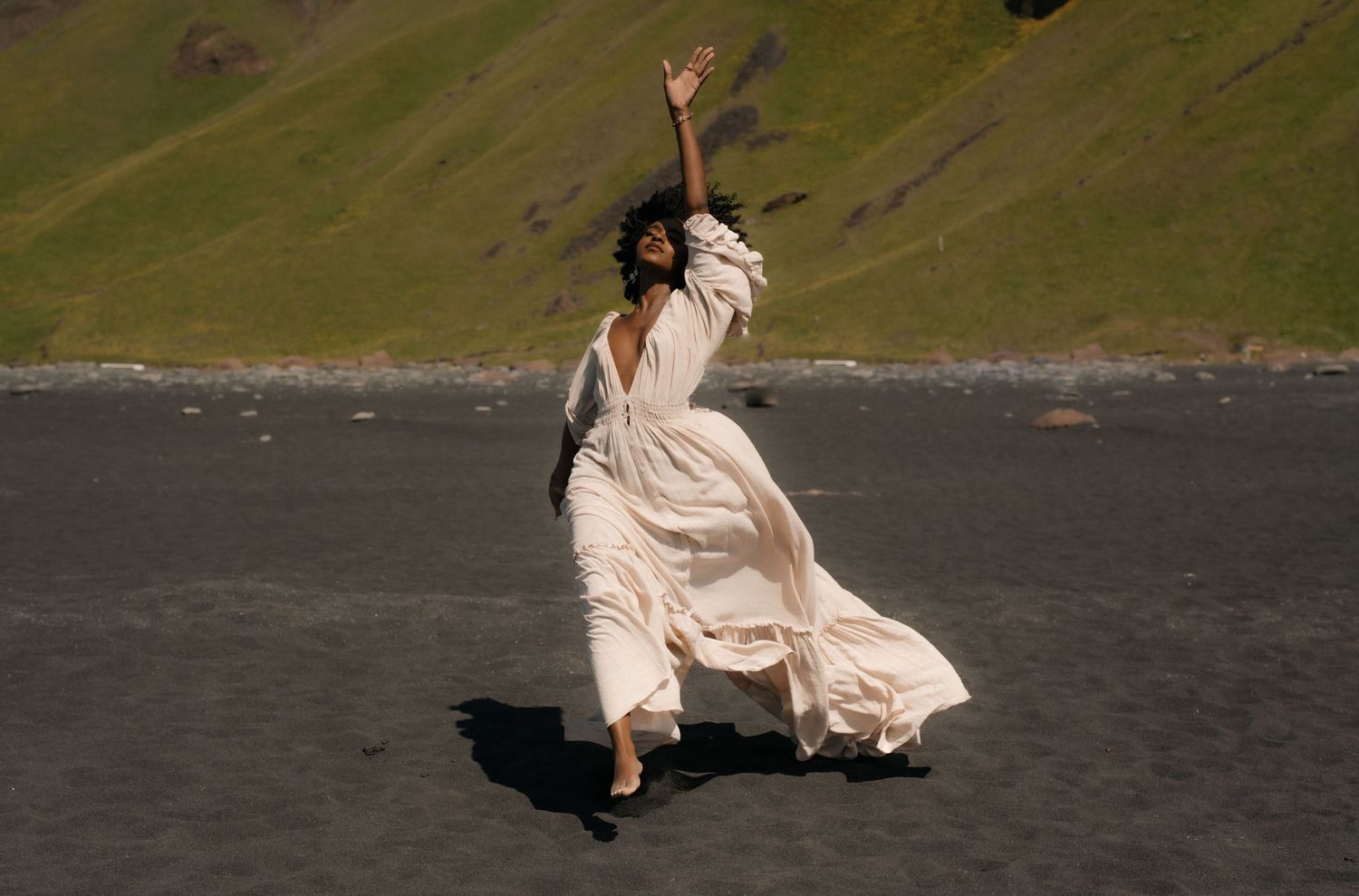 A woman in a flowing white dress dances gracefully on a black sand beach with green hills behind her.