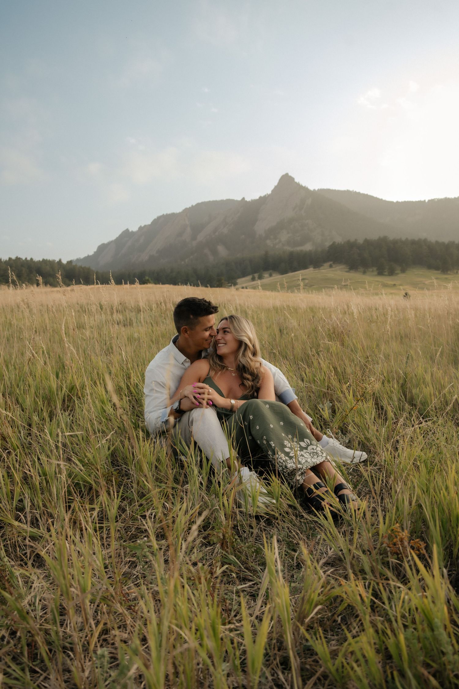 Couple sitting in tall grass field with Rocky Mountain Flatirons backdrop during golden hour engagement photo session.