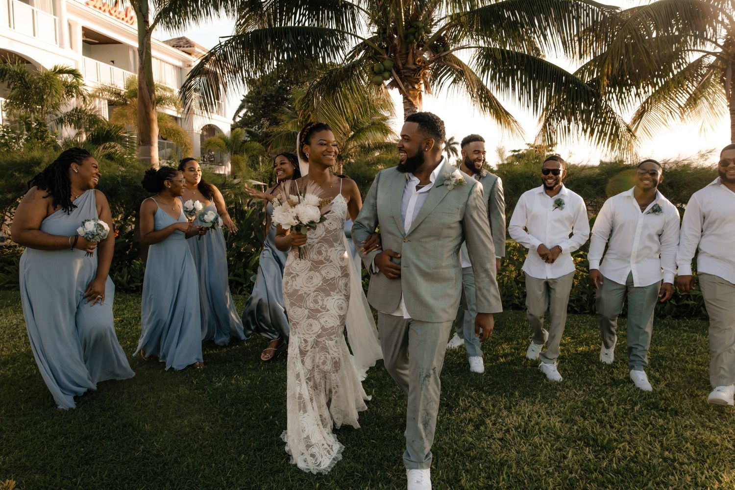 Wedding party in light blue dresses and white outfits poses on grass with palm trees at sunset.