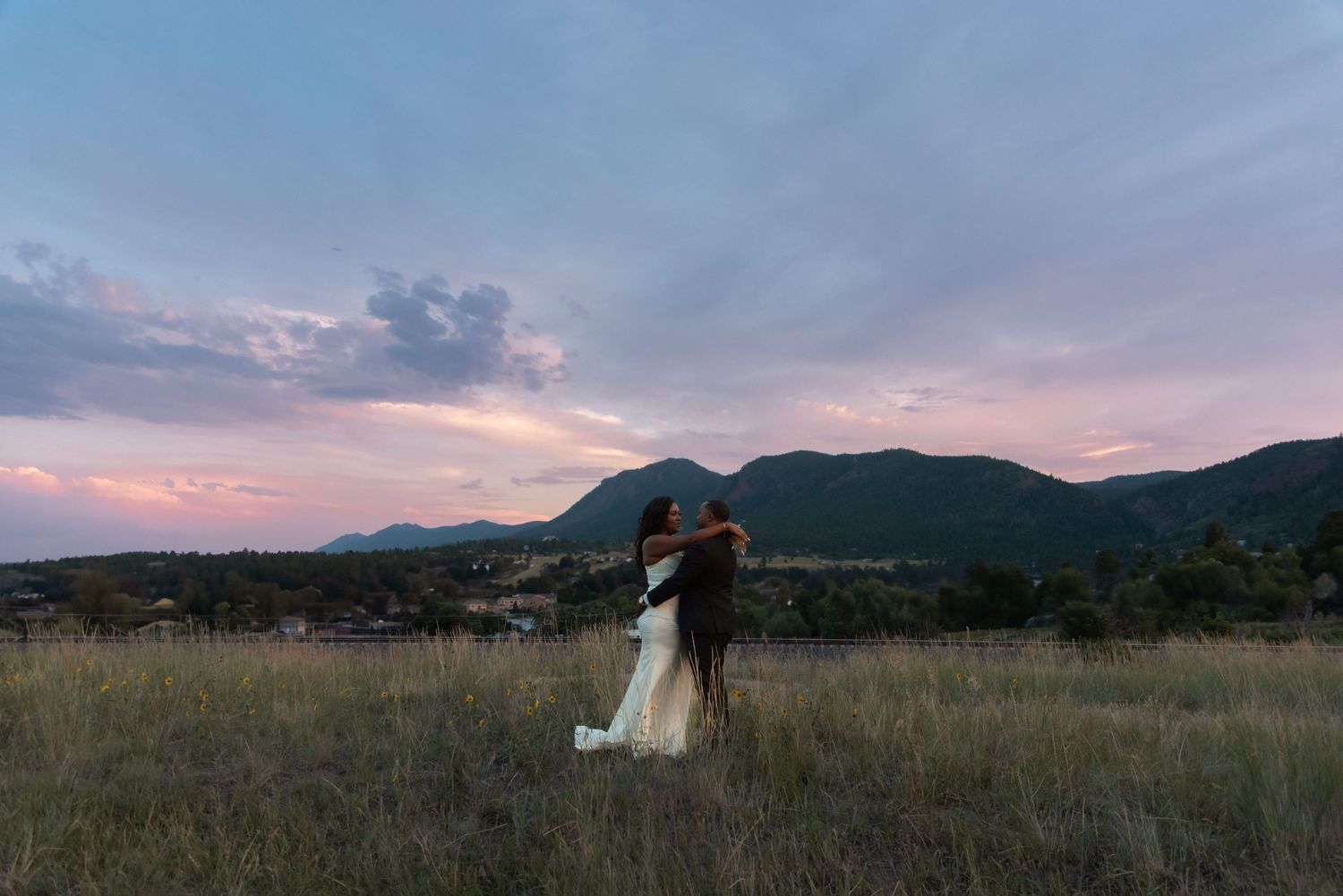 Couple embracing in a golden field at sunset with mountain backdrop and colorful pink and blue sky.