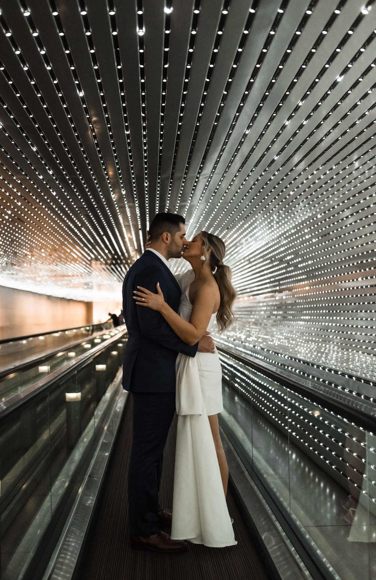 A couple shares a romantic kiss on an illuminated moving walkway tunnel with sparkling light patterns creating a magical effect.
