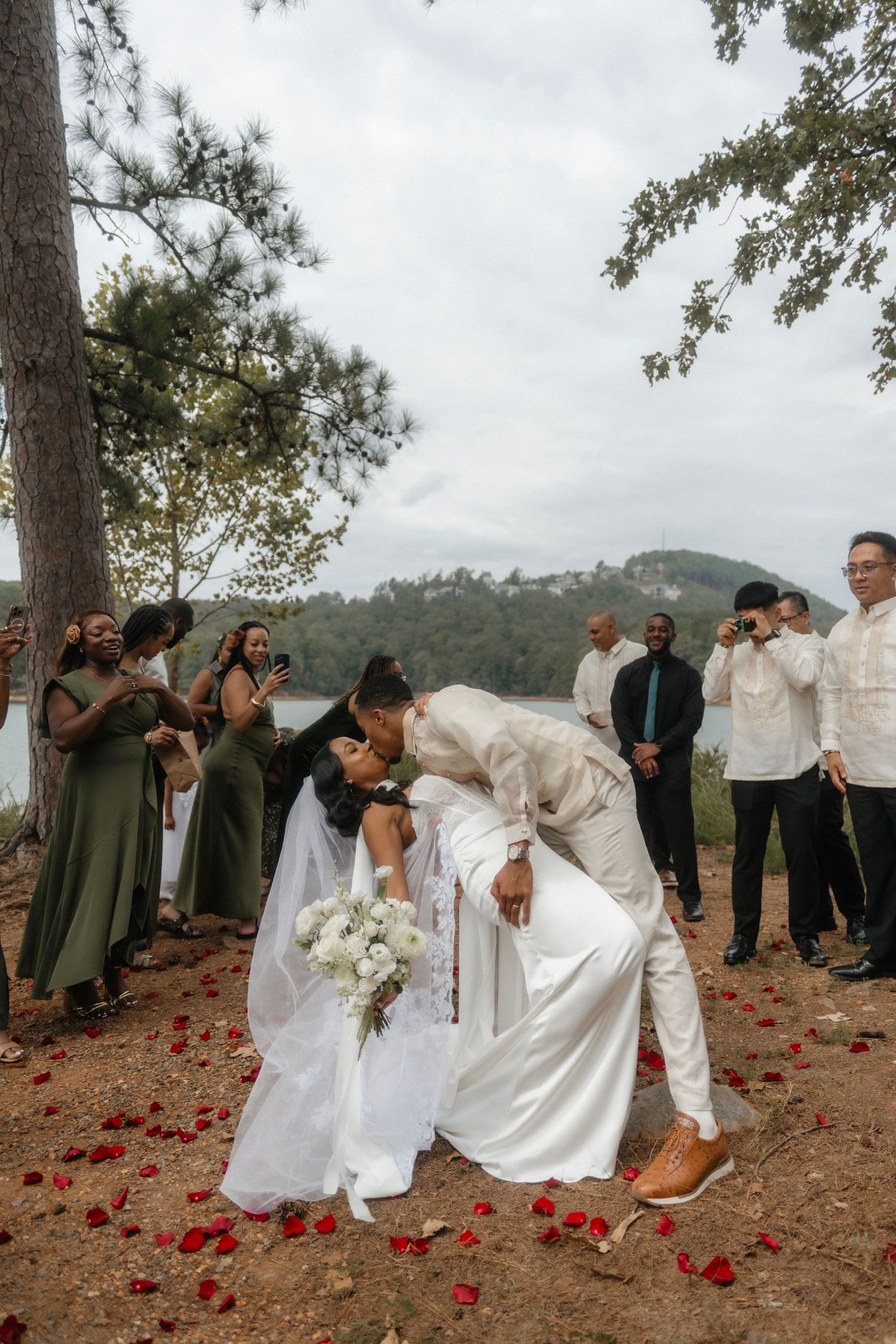 Couple sharing a romantic first kiss at outdoor wedding ceremony with bridal party, rose petals, and lake backdrop.