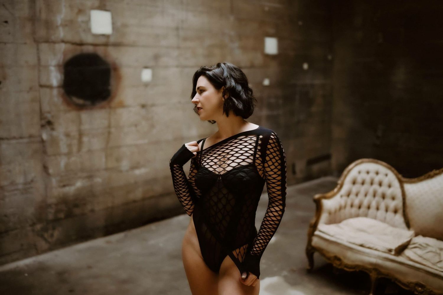 A figure in a black fishnet bodysuit poses dramatically against a rustic concrete wall with an antique couch in the background.