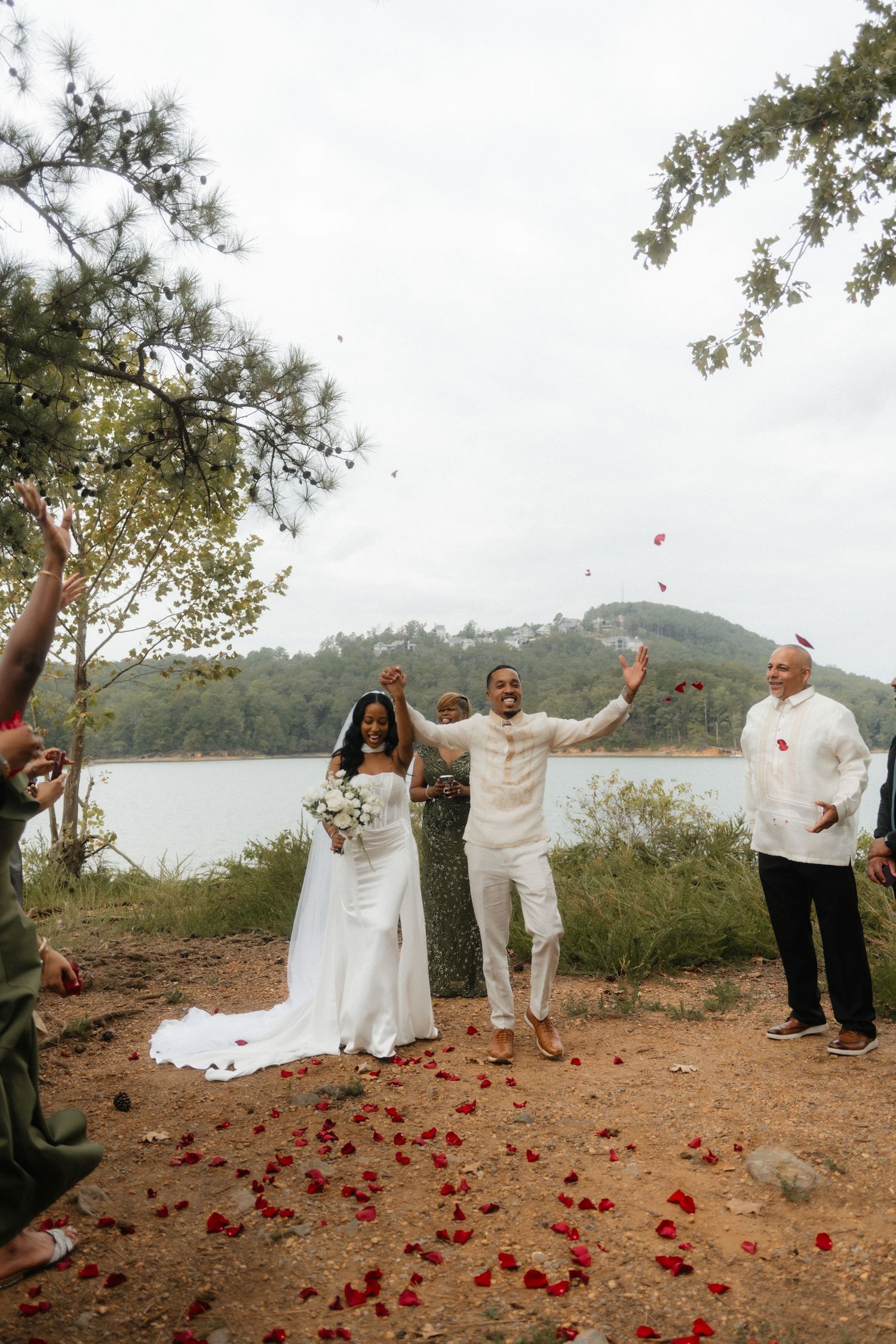 Couple in white celebrates wedding by lakeside with rose petals, officiant, and guests cheering outdoors.