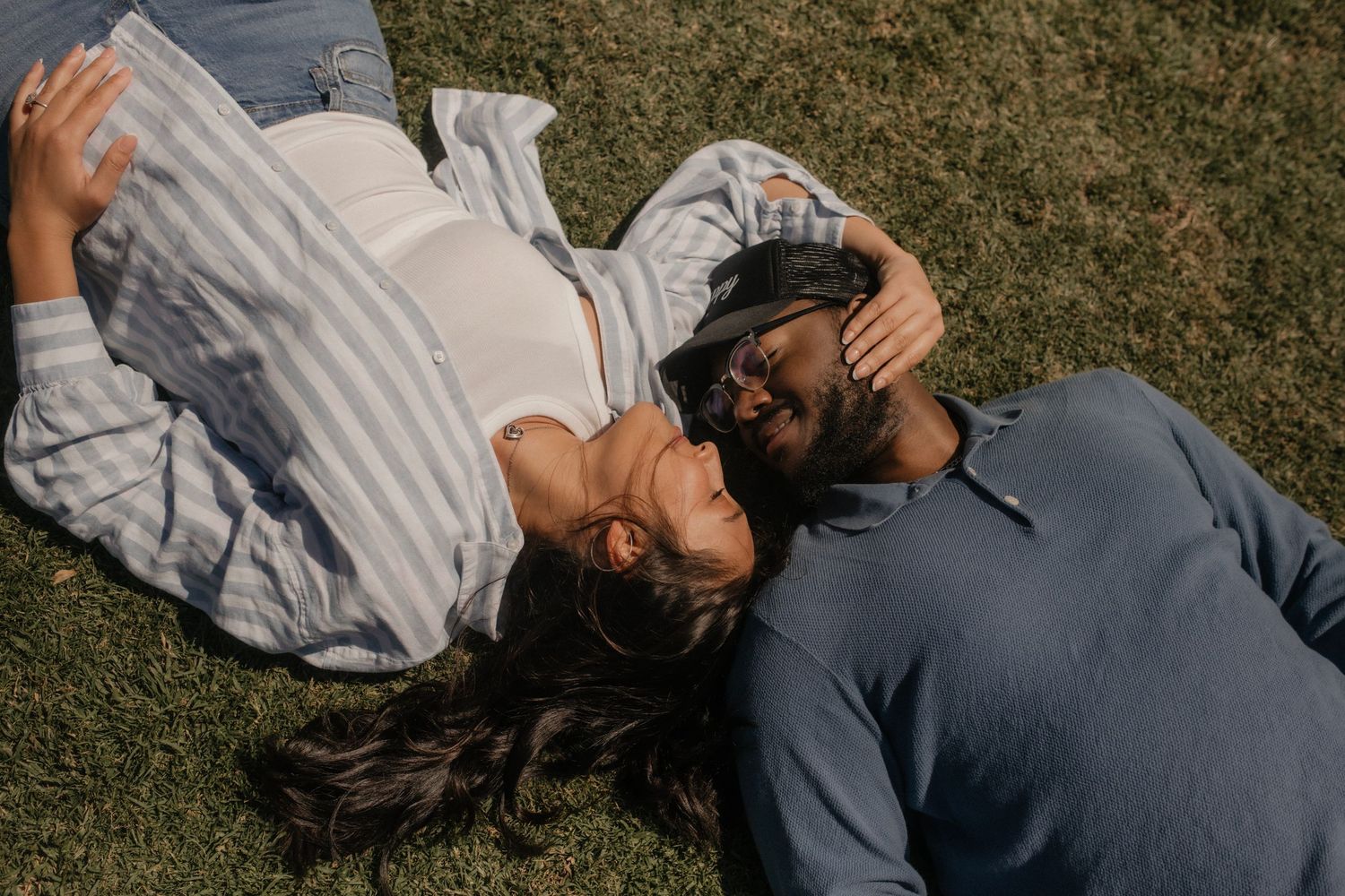 A couple lying on green grass, facing each other intimately, wearing casual shirts on a sunny day.