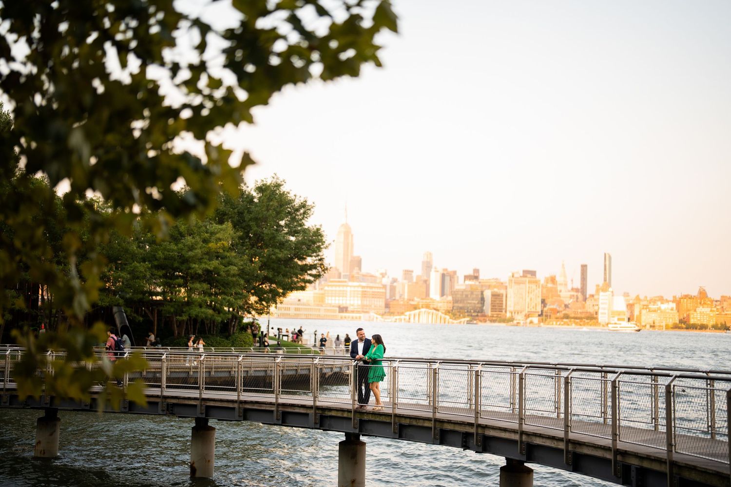 Scenic waterfront boardwalk with city skyline and trees in background at sunset.