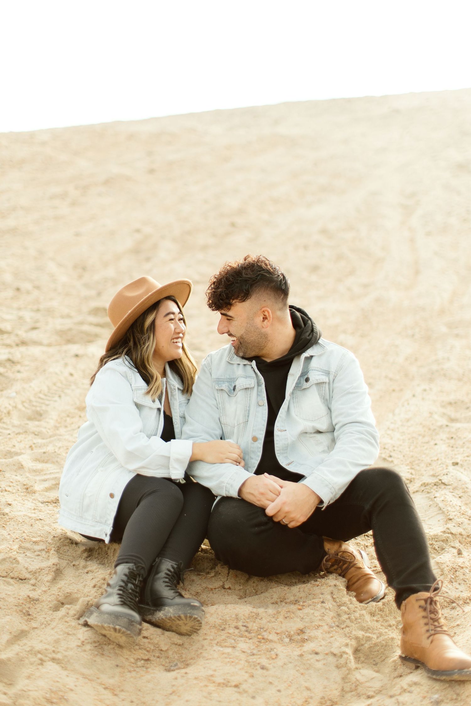 Couple in light denim jackets shares intimate moments while sitting on sandy beach.