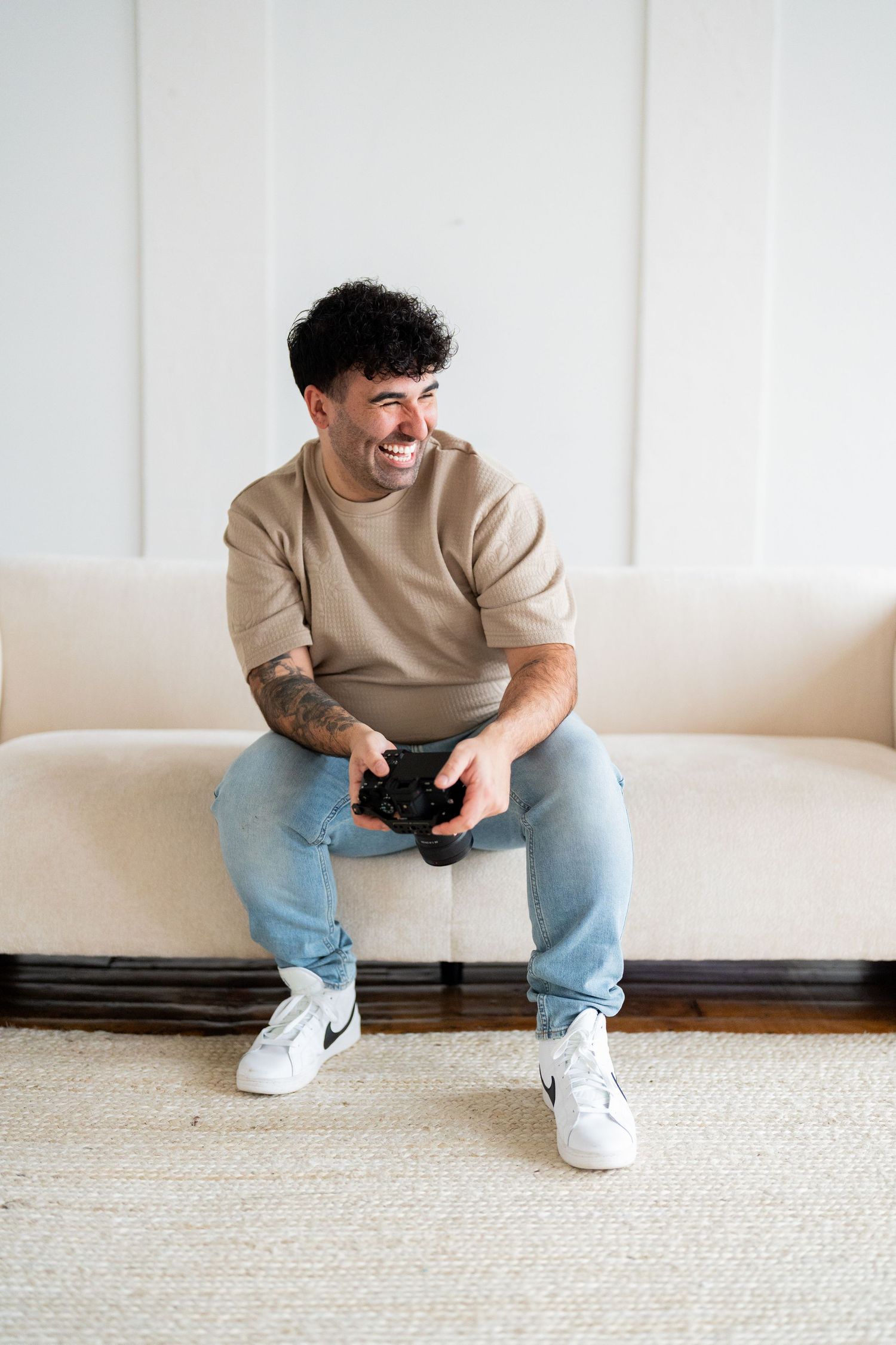 Person in beige sweater and jeans sits casually on white carpet against minimalist wall.