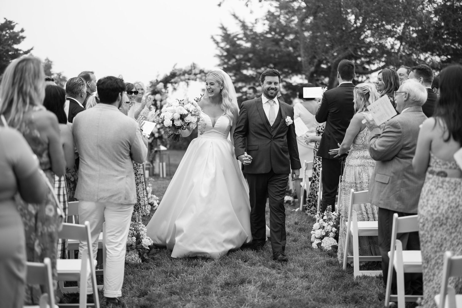 Wedding guests gathered outdoors for ceremony with white chairs arranged on lawn.