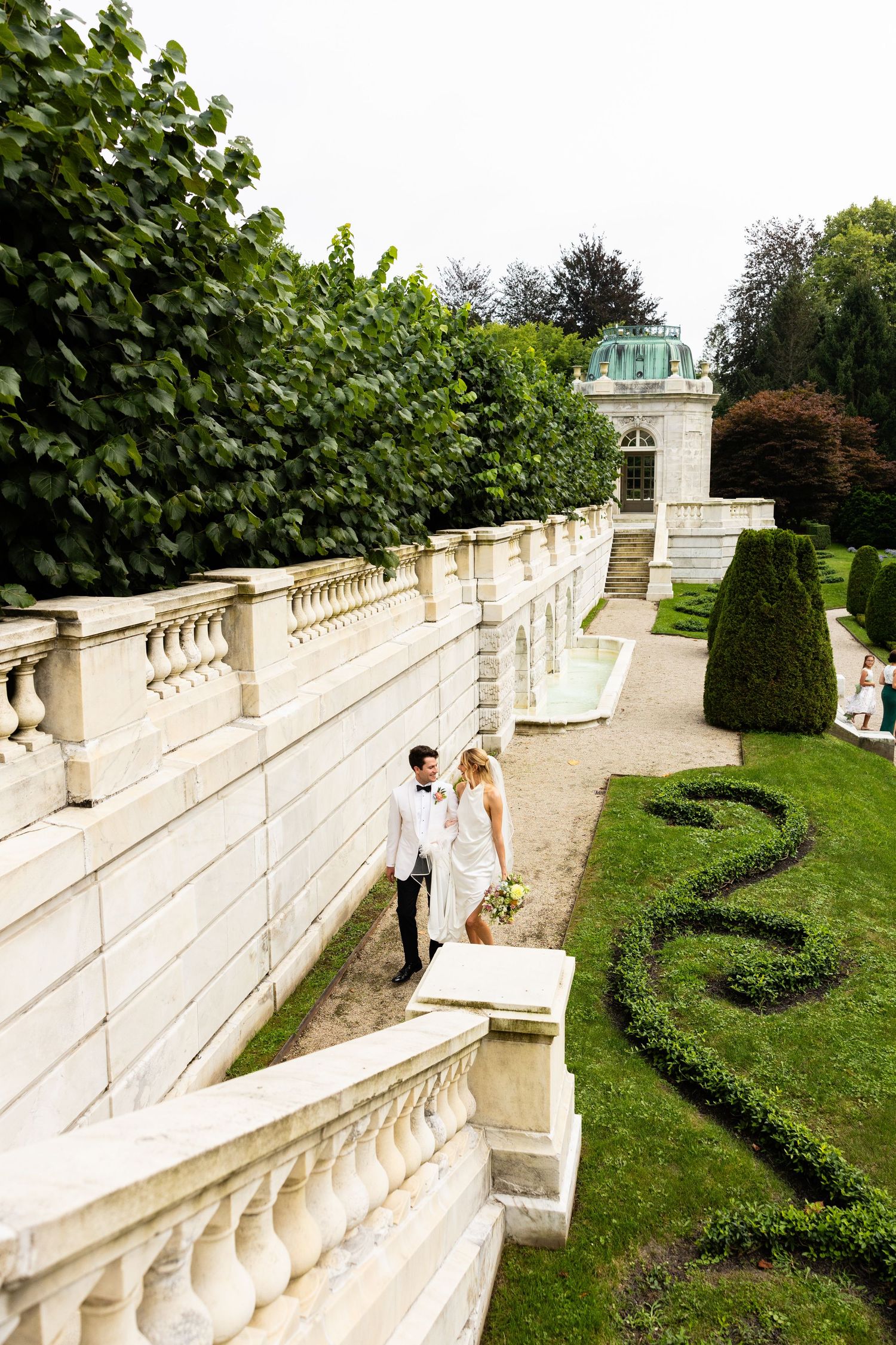 A couple shares an intimate moment on ornate garden steps with manicured hedges.