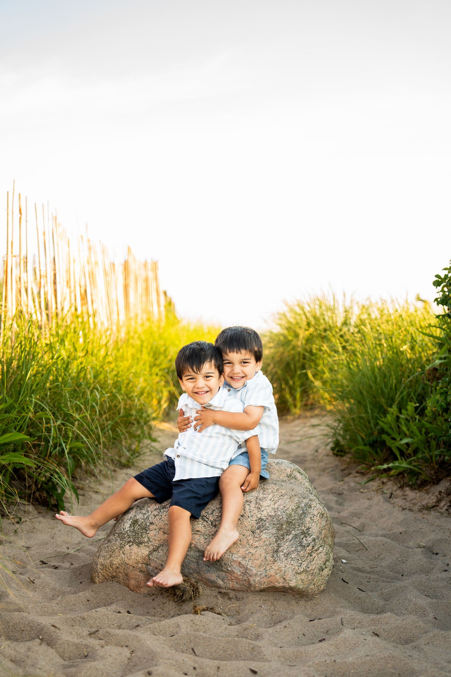 Two children sit together on a large rock at the beach with tall grass in the background.