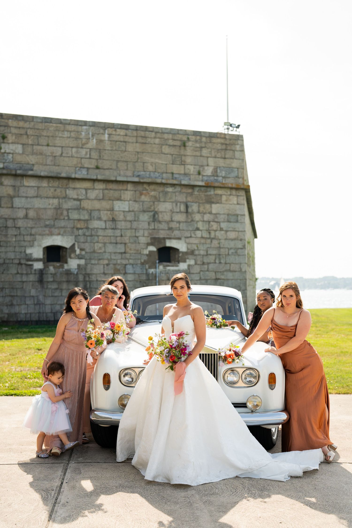 Wedding party poses with classic white car in front of stone fortress by waterfront.