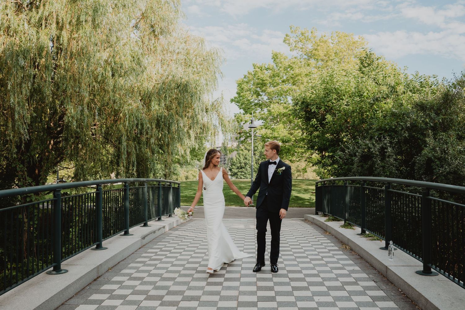 A couple walks hand in hand across a patterned bridge surrounded by greenery.