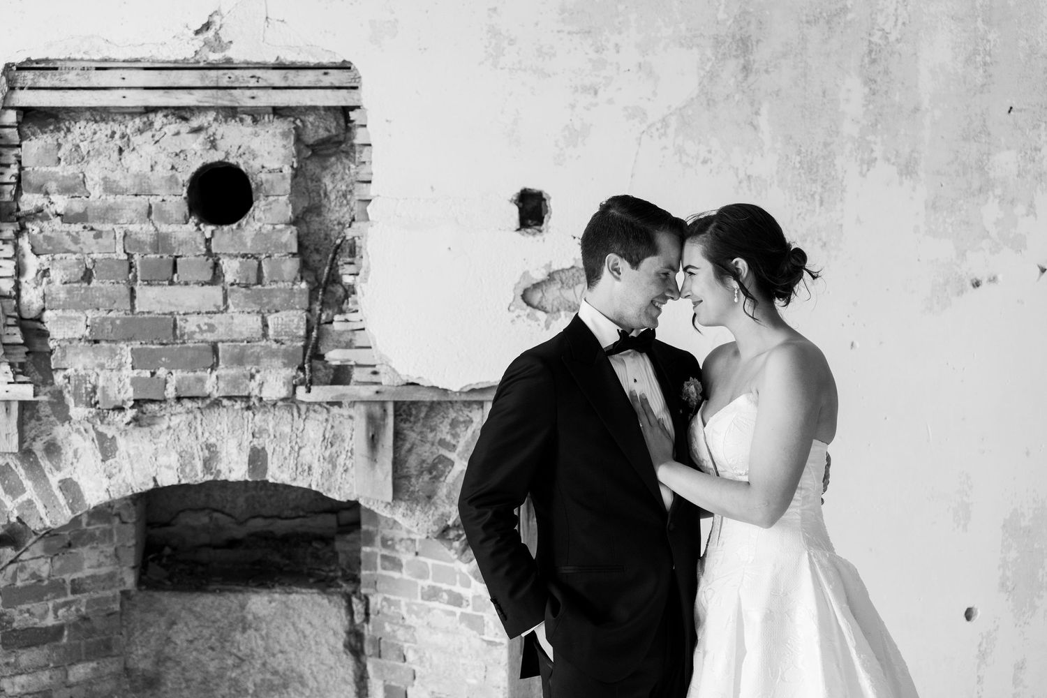 Black and white wedding photo of bride and groom sharing intimate moment by rustic brick fireplace.