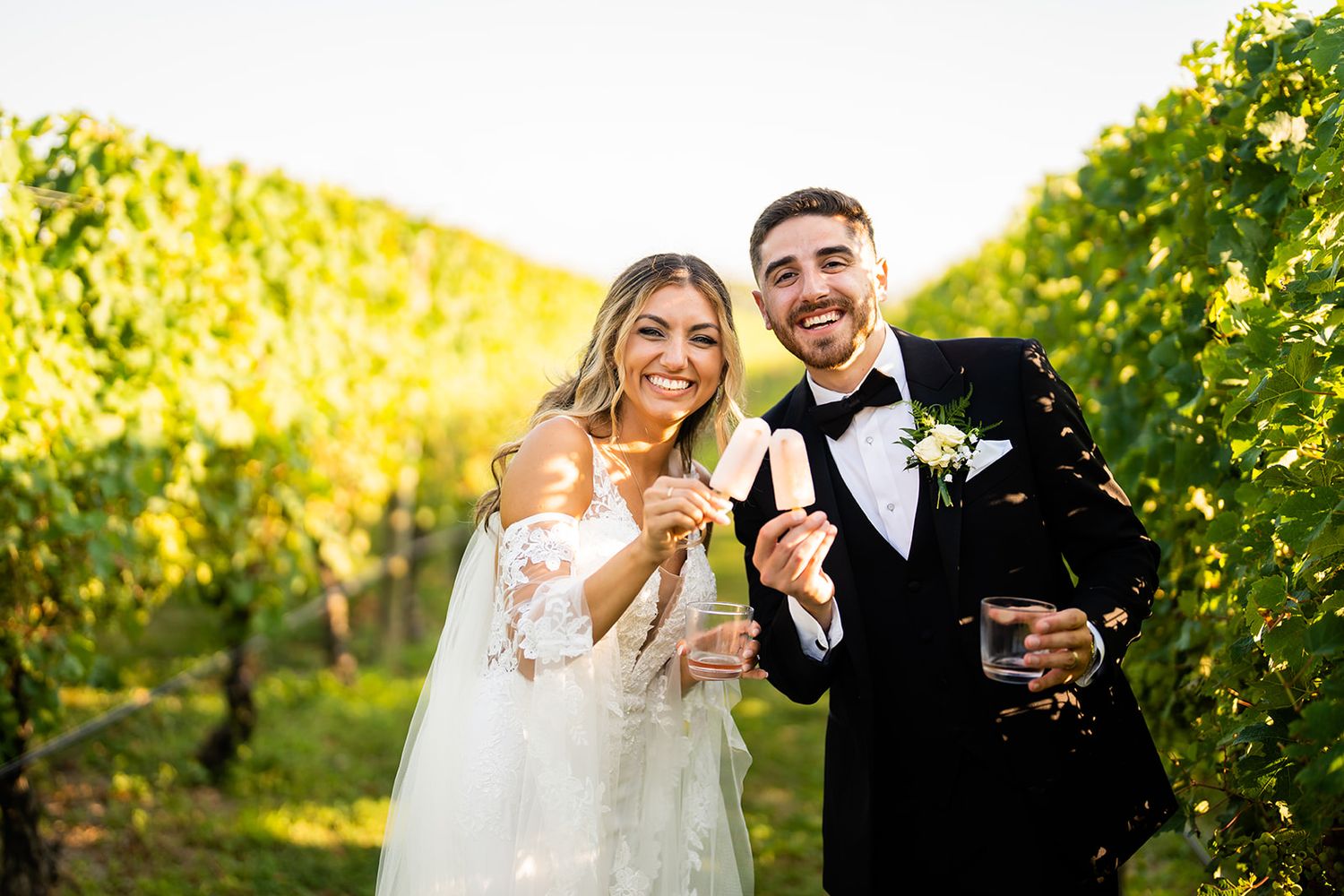 Newlywed couple sharing champagne toast among sun-lit vineyard rows during golden hour celebration.