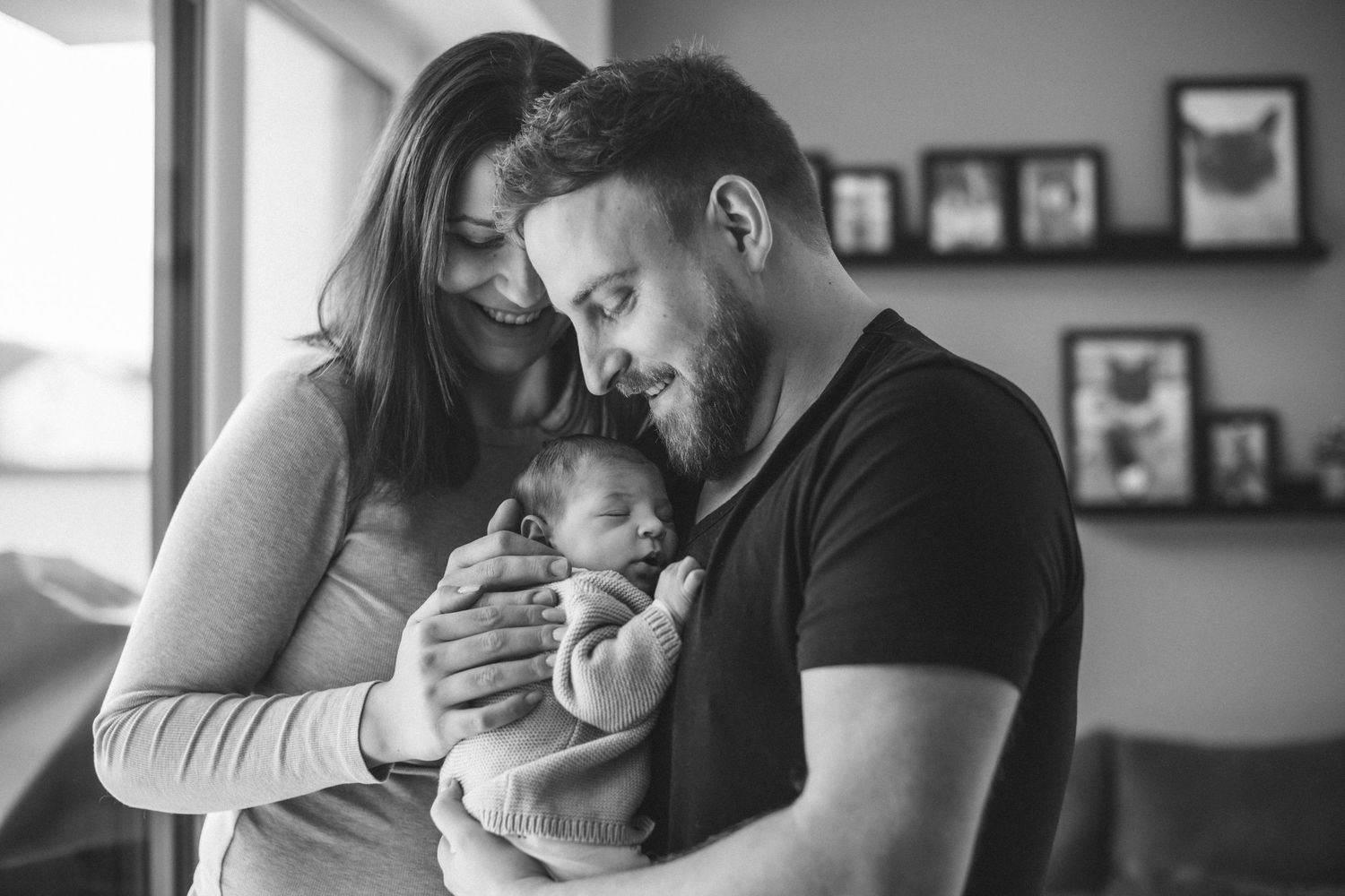 Intimate black and white portrait of new parents tenderly holding their newborn baby indoors.