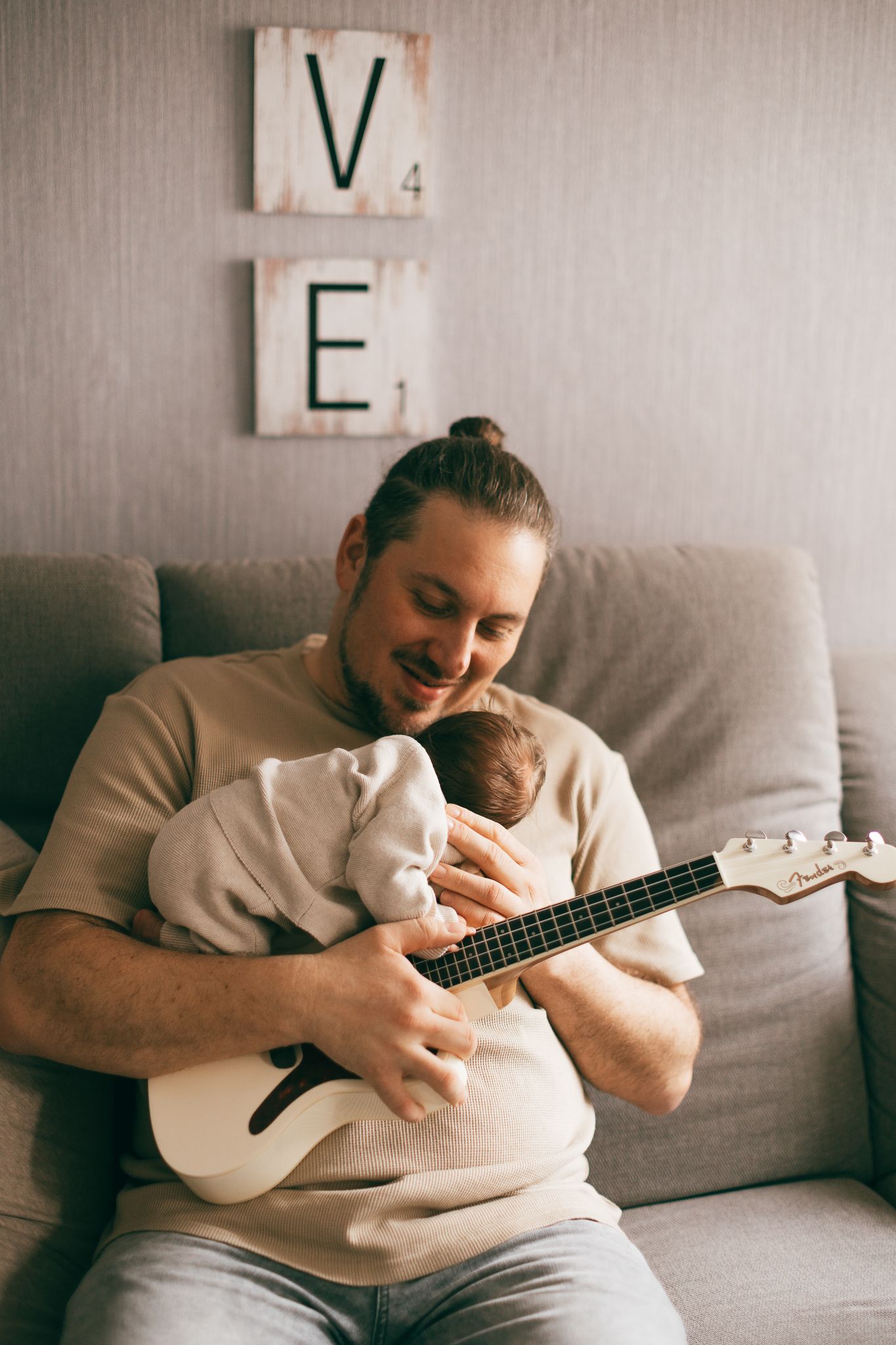 Papa hält und spielt eine kleine Ukulele mit seinem Baby auf einem grauen Sofa in Konz.