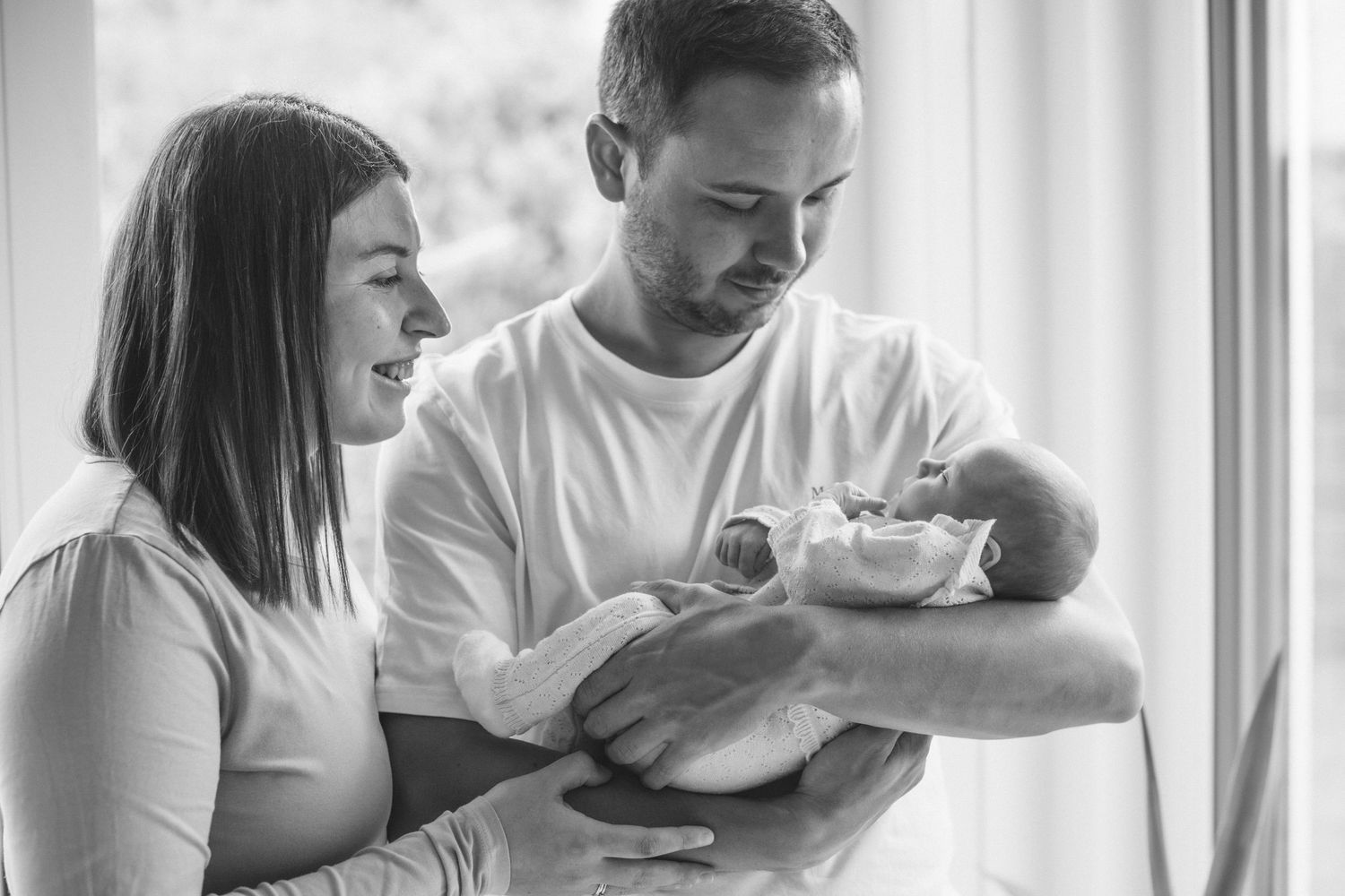 A tender family moment as new parents gaze lovingly at their newborn baby by a bright window.