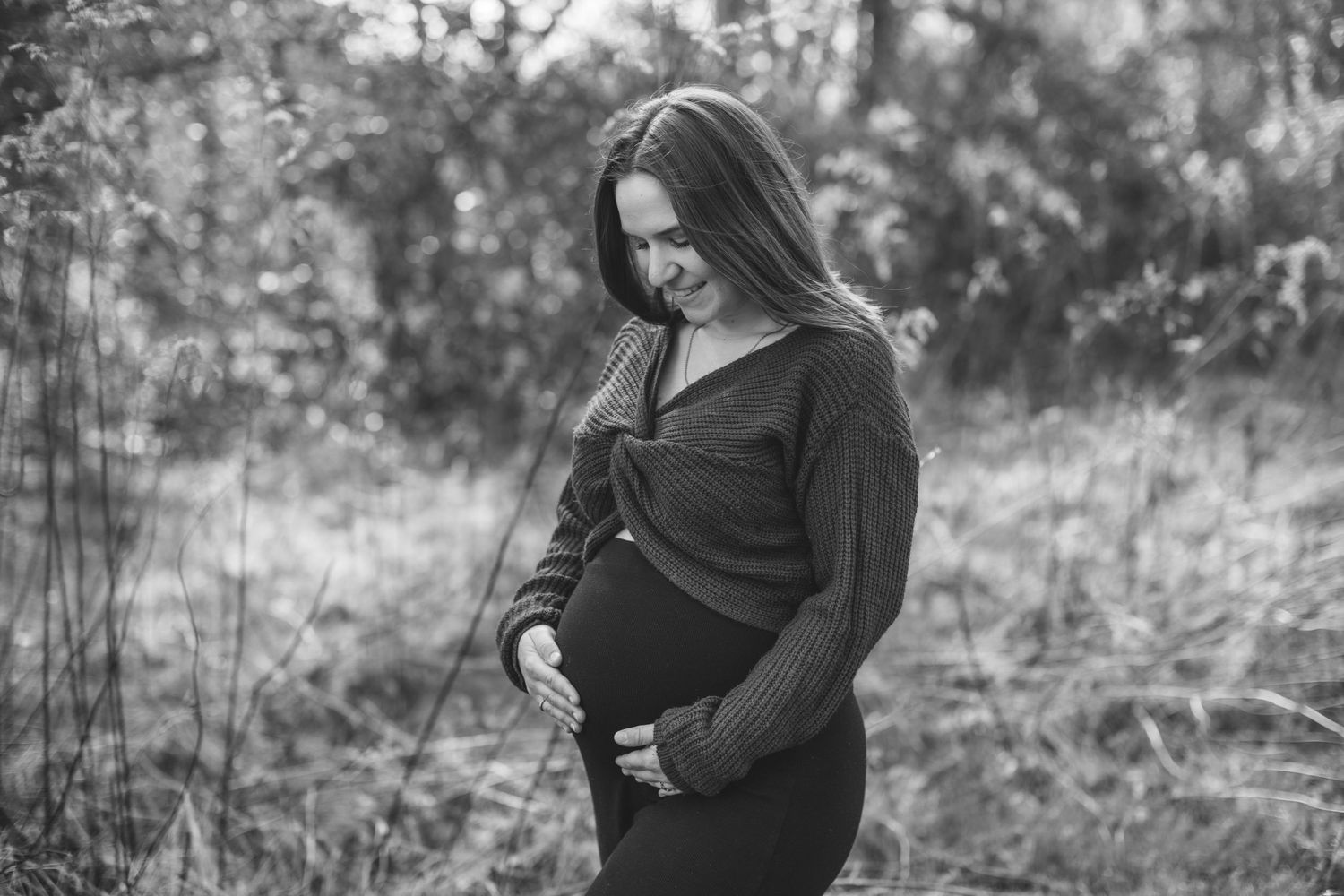 Expectant woman in black dress caresses her pregnant belly in a serene forest setting.
