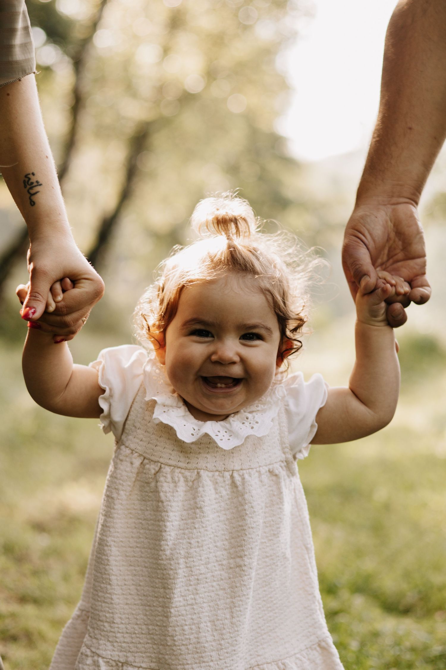 Smiling baby in white lace dress being held up by parent's hands against blurred outdoor background.