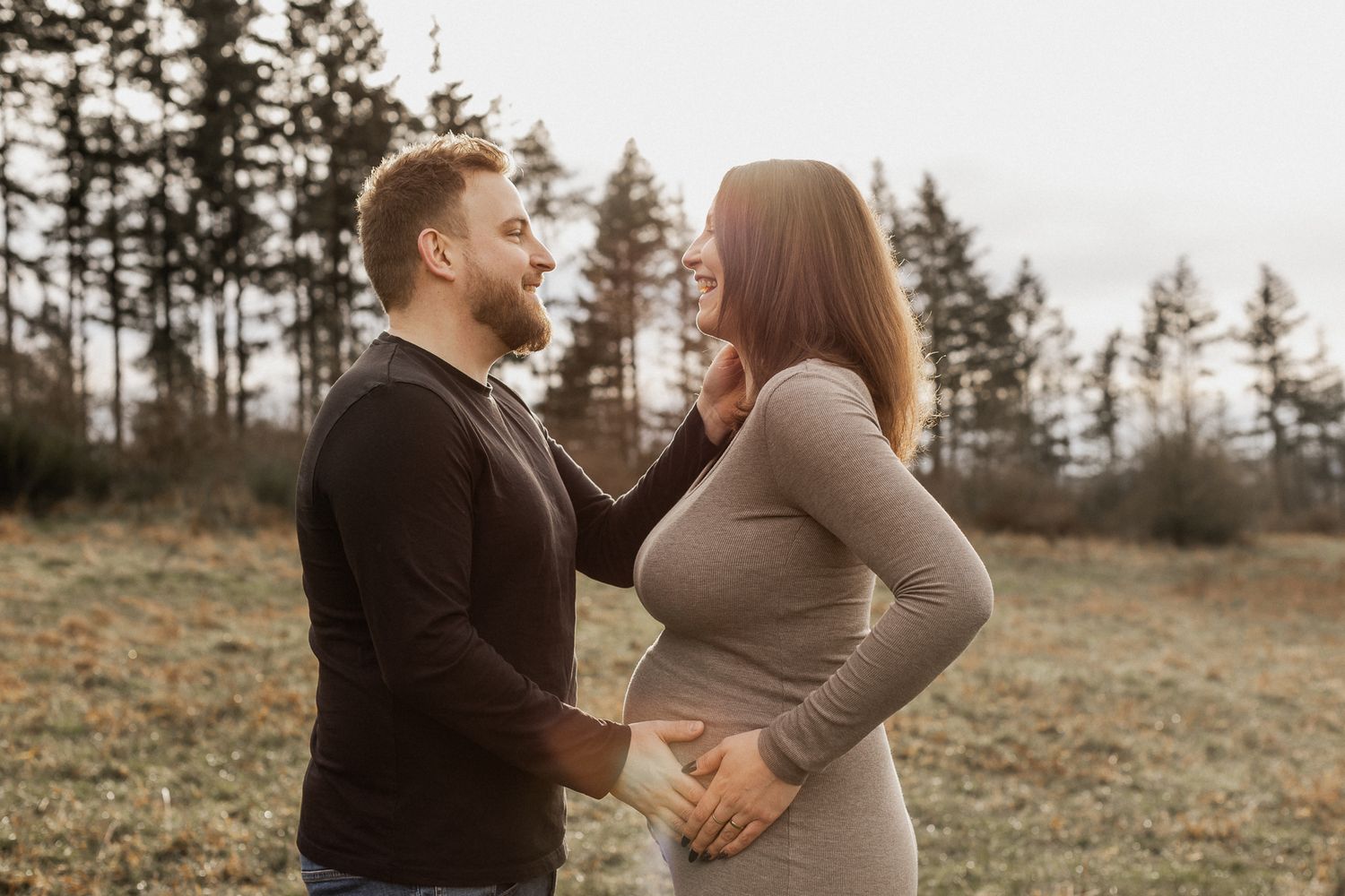 Ein Paar teilt einen emotionalen Moment auf einem Feld im Freien, mit Babybauch und immergrünen Bäumen im Hintergrund.