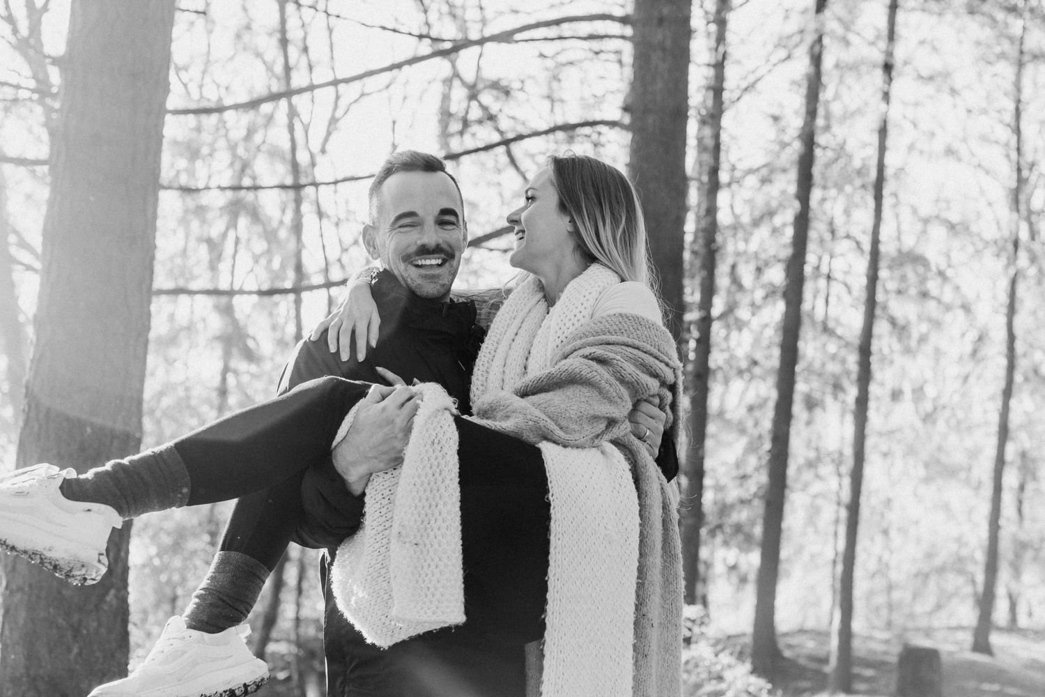 A couple shares joyful laughter while wrapped in blankets against a backdrop of tall forest trees.
