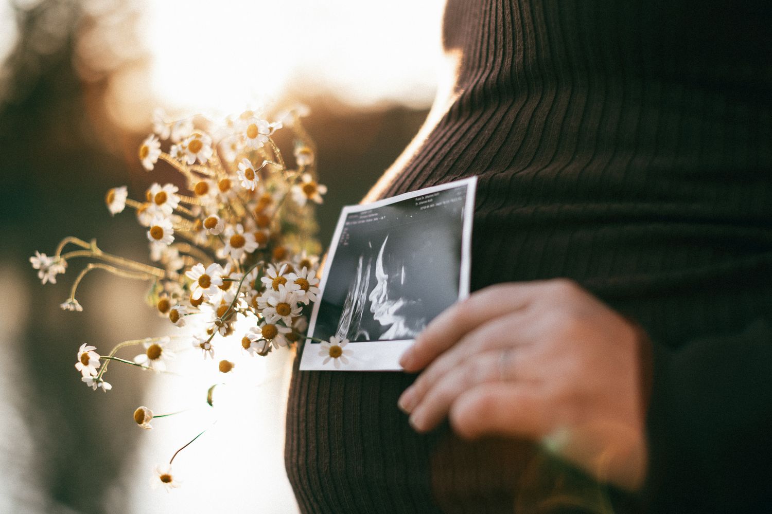 Eine Person hält ein Ultraschallbild an den Babybauch, umgeben von getrockneten Blumen im Licht des Sonnenuntergangs in Konz.