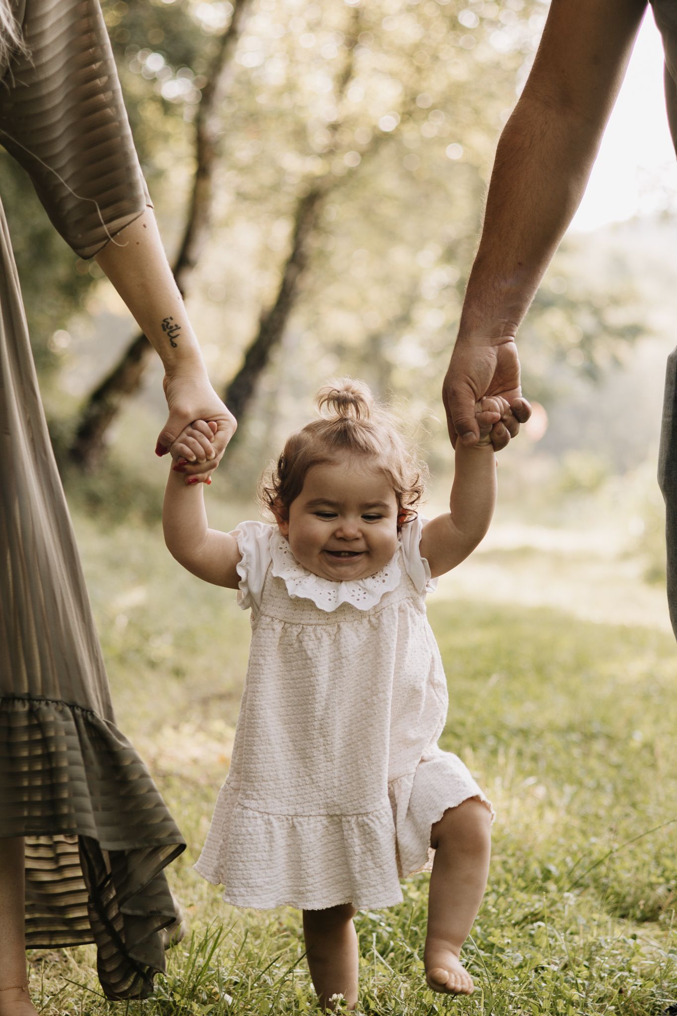 Ein Baby in einem weißen Kleid macht seine ersten Schritte, liebevoll gestützt von helfenden Händen – inmitten einer Naturkulisse in Konz.