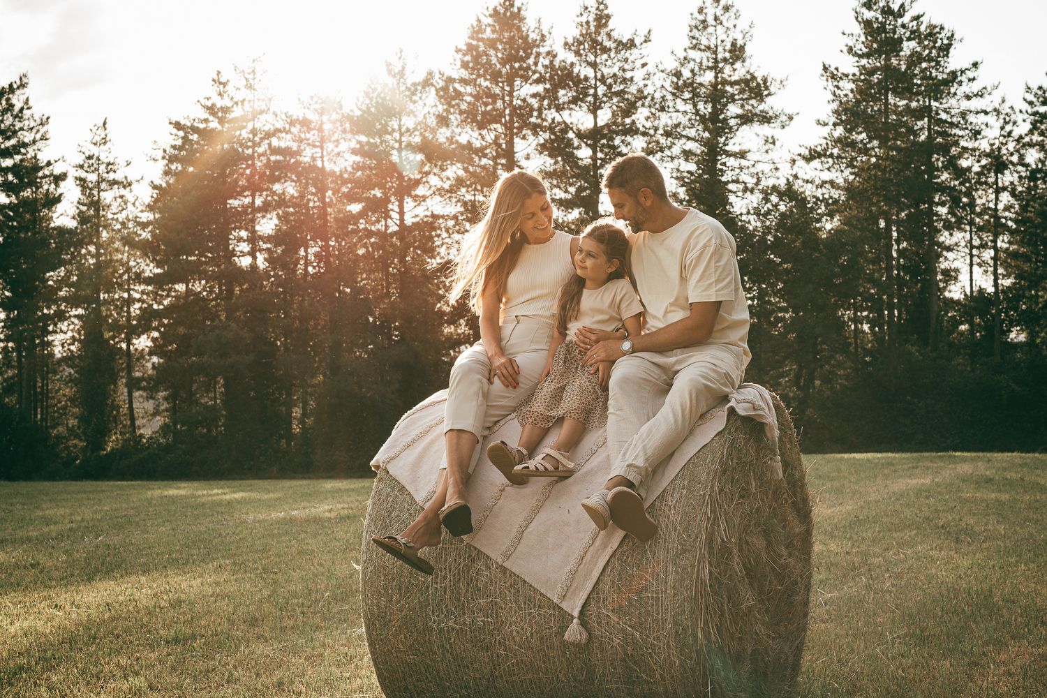 Familie sitzt gemeinsam auf einem Heuballen in einem von der Abendsonne beleuchteten Feld mit Kiefern. Familienfotografie